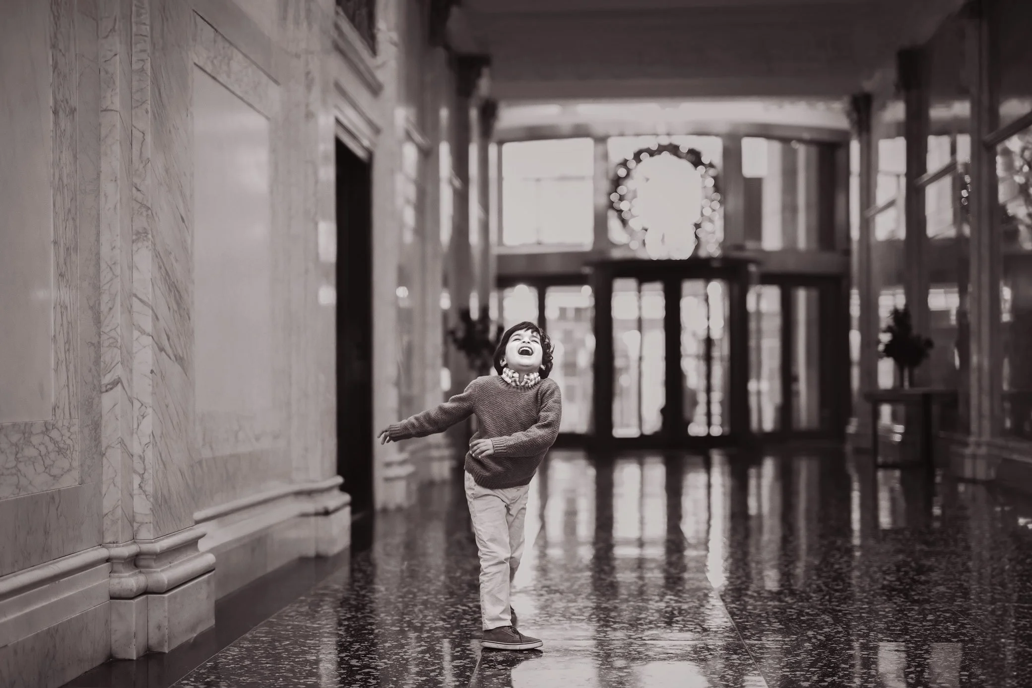 A young boy with curly hair laughing and playing in a grand, well-lit hallway with marble walls and a reflective floor, decorated with a Christmas wreath in the background.
