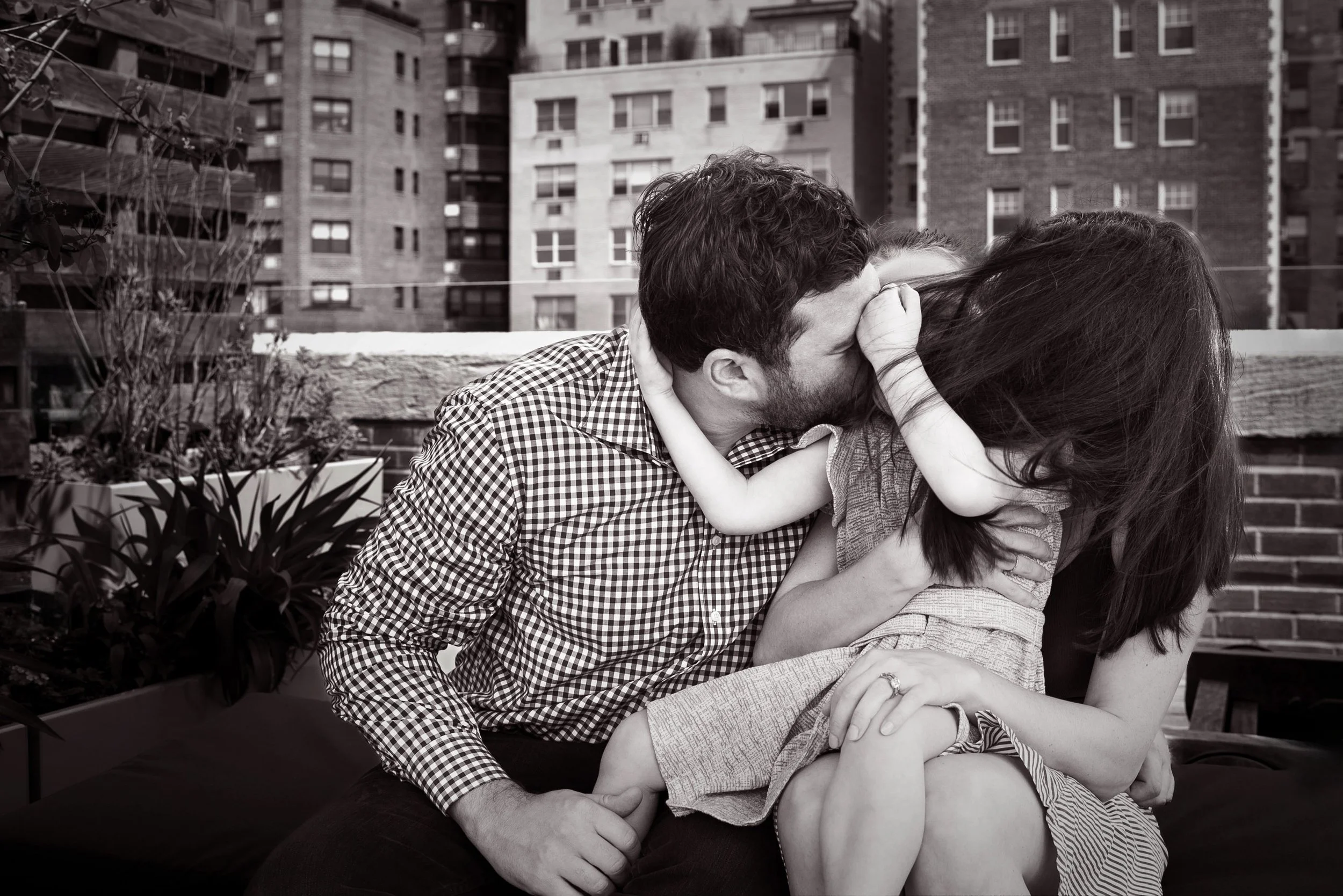 A black and white photograph of a couple sitting outdoors on a rooftop or balcony, sharing an intimate moment of kissing. The man has curly hair and a beard, wearing a checkered shirt, while the woman has long dark hair and is wearing a light-colored