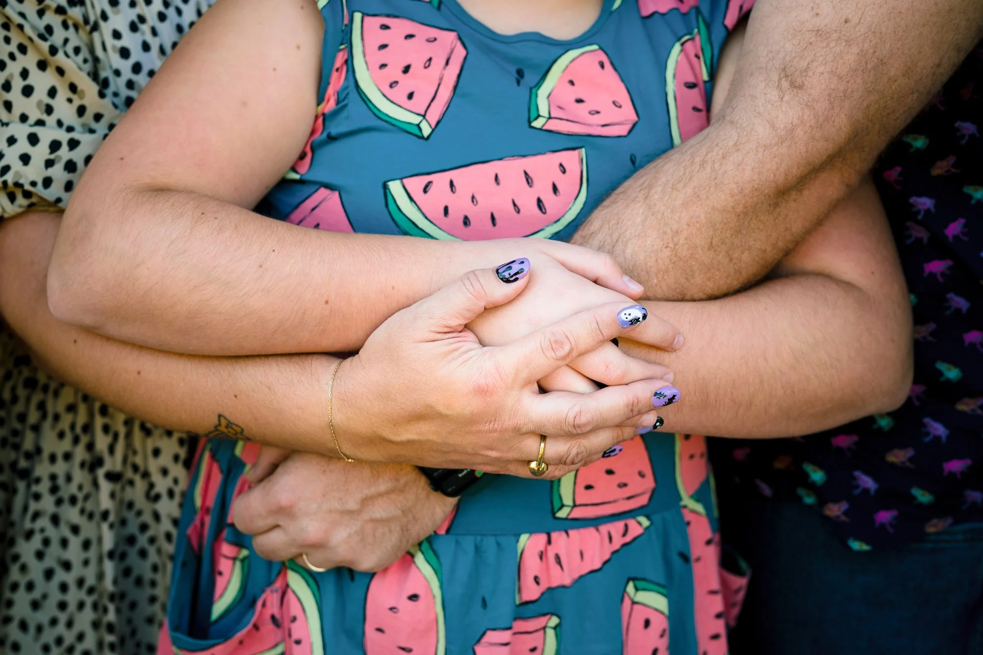 Close-up of a person’s arm with a gold bracelet and ring, holding hands with another person, wearing a shirt with a watermelon pattern, surrounded by people with various patterned clothing