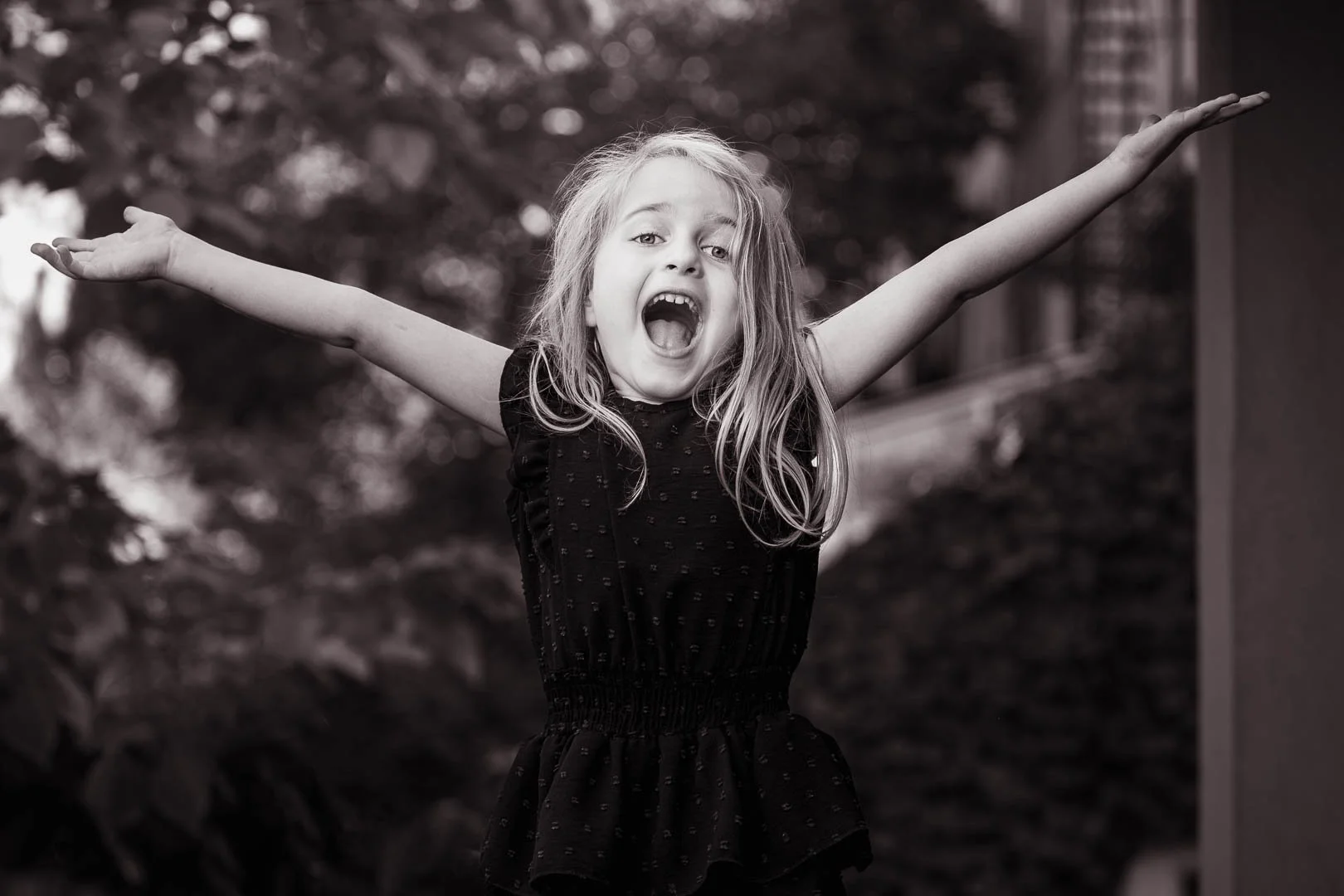 A young girl with long hair, wearing a dark dress, with her arms outstretched and her mouth open in a joyful expression outdoors.