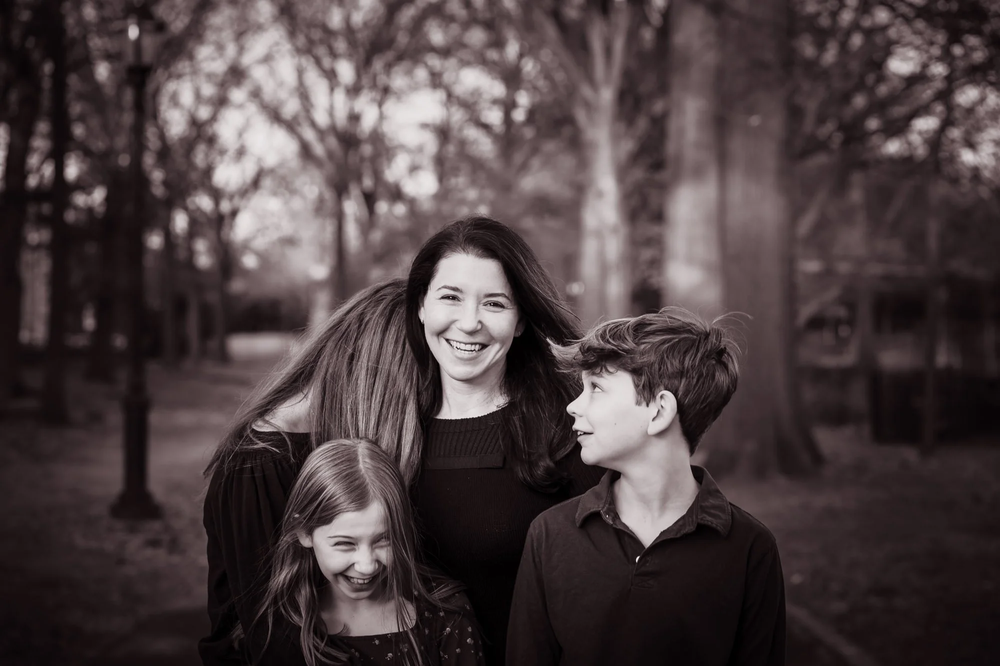 Black and white photo of a woman with long hair smiling, surrounded by three children, in a park with trees in the background.