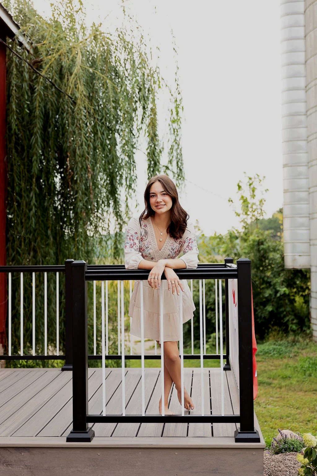 A woman standing on a wooden porch railing outdoors, smiling, with greenery and trees in the background.