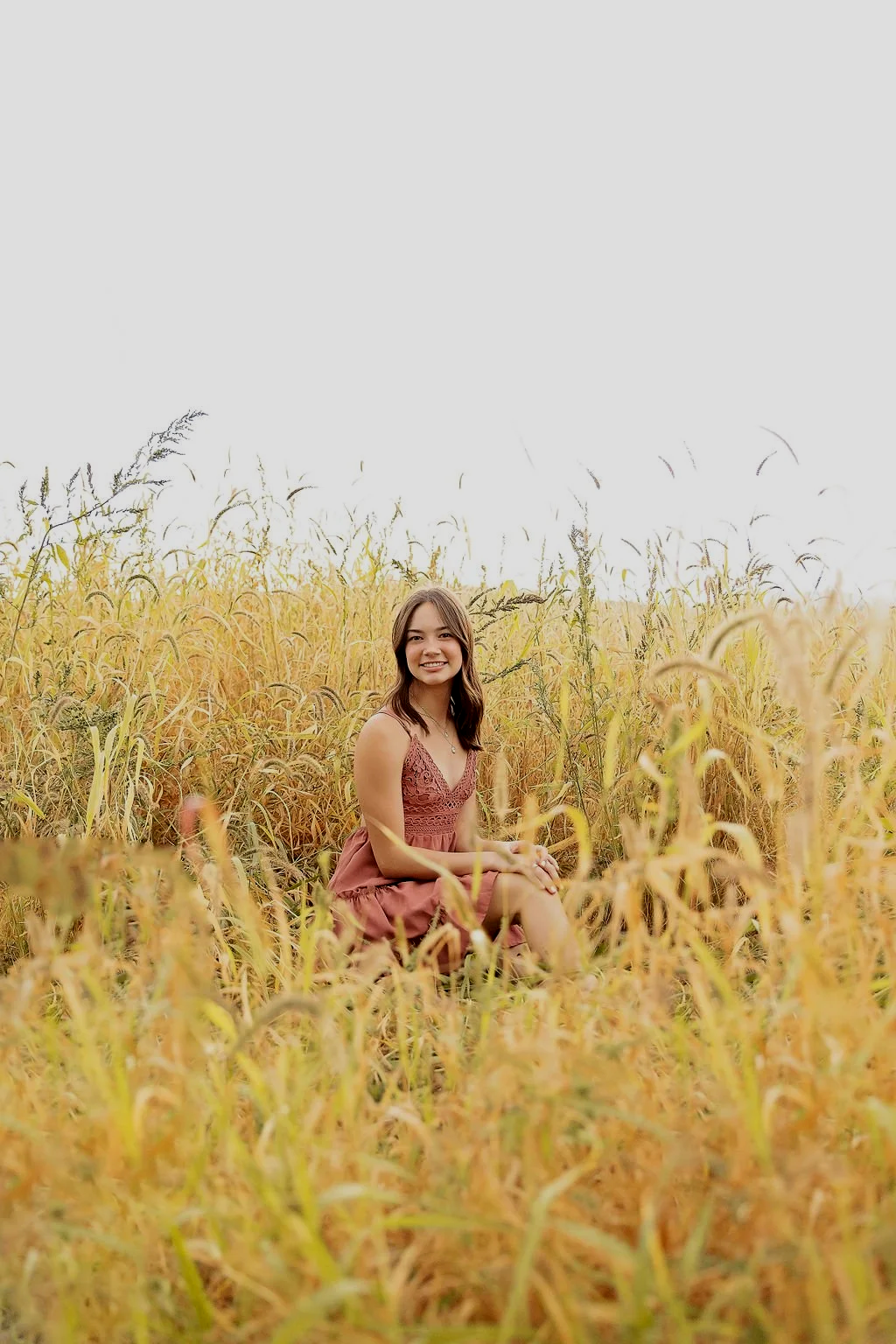 A young woman in a mauve dress sitting in a field of tall, yellow grass, smiling at the camera.