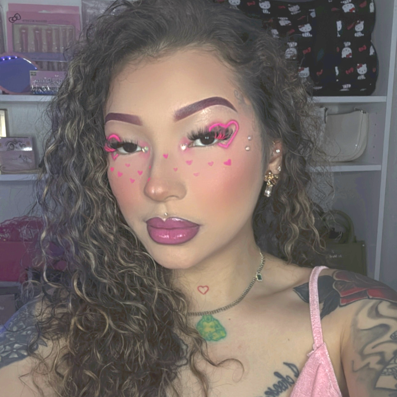 A woman with long, curly hair and multiple facial piercings taking a selfie. She has pink makeup, heart-shaped face paint, and is wearing a pink top with a tattoo on her shoulder. She also has necklaces and earrings, and there are shelves with various items in the background.