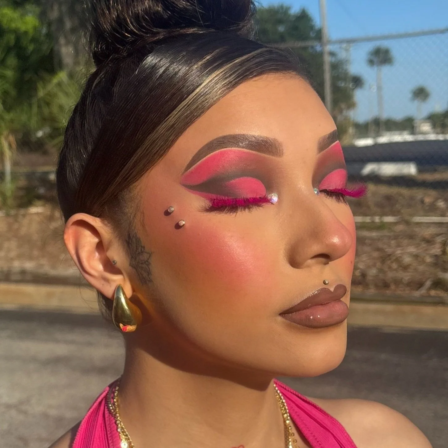 A woman with styled dark hair, bold pink and black eye makeup, pink false eyelashes, gold earrings, and a pink top, with a blurred outdoor background.