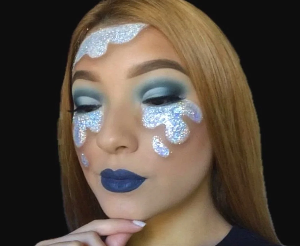 A woman with long, straight red hair and makeup featuring blue eyeshadow, dark blue lipstick, and glitter tears on her face, set against a black background.