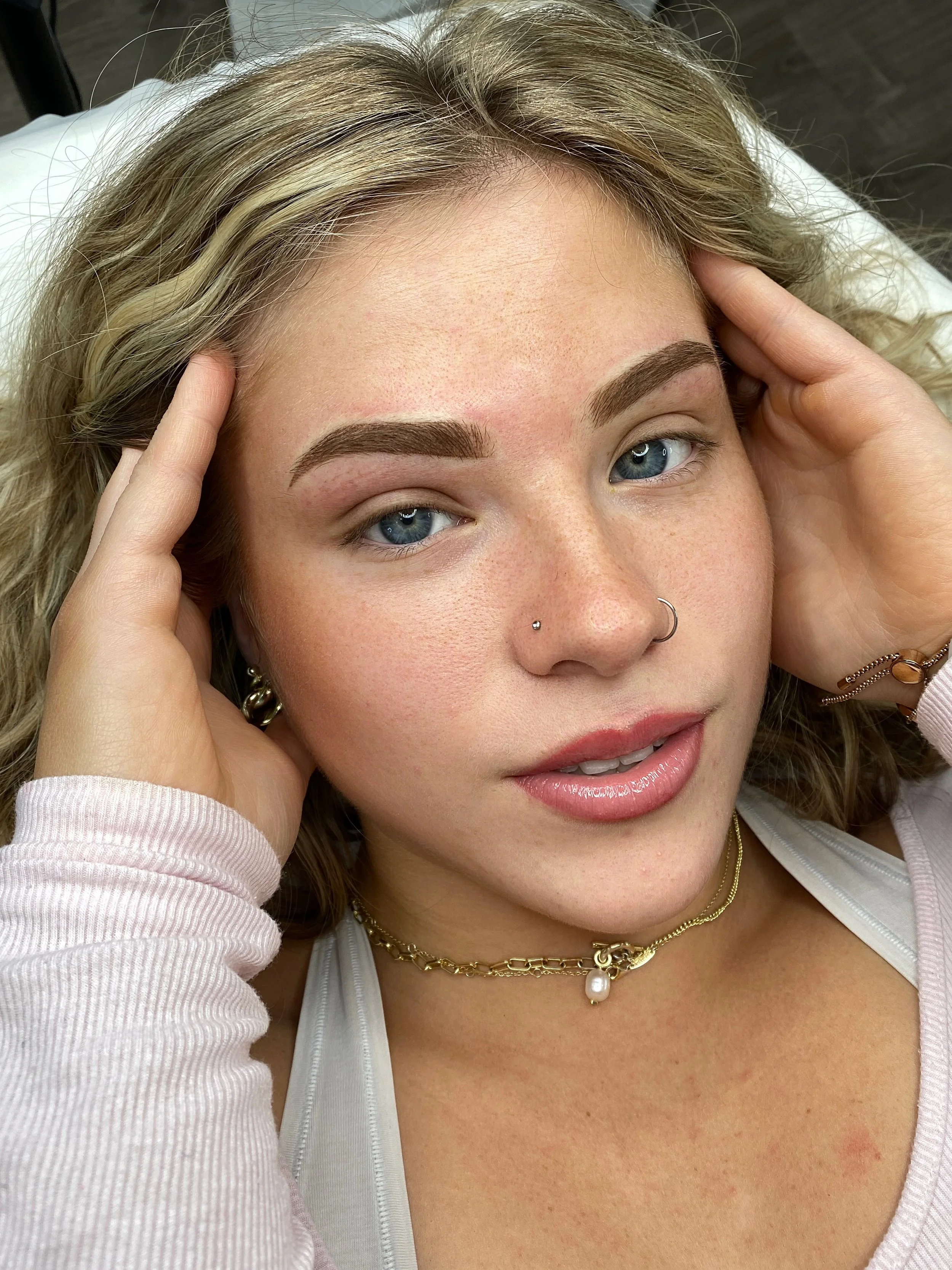 Close-up of a woman with blonde curly hair, blue eyes, and multiple piercings, lying on a bed with her hands touching her head, wearing jewelry and a white top.