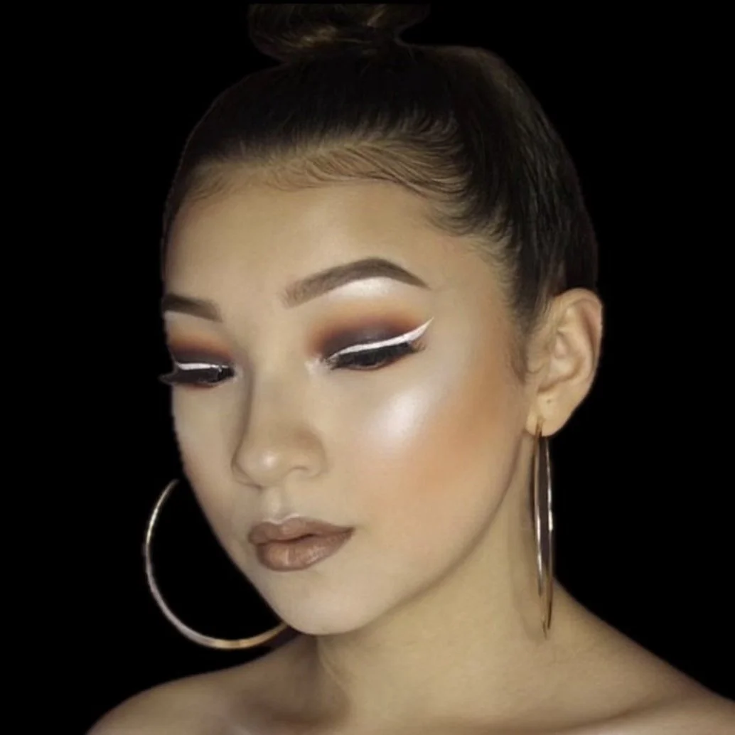 Close-up of a woman with makeup, including eyeshadow, eyeliner, and lipstick, wearing large hoop earrings, against a black background.