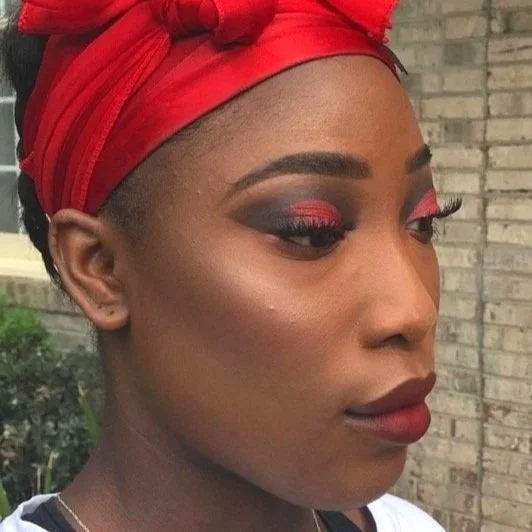 Close-up of a woman with vibrant makeup, red headscarf, and a brick wall background.