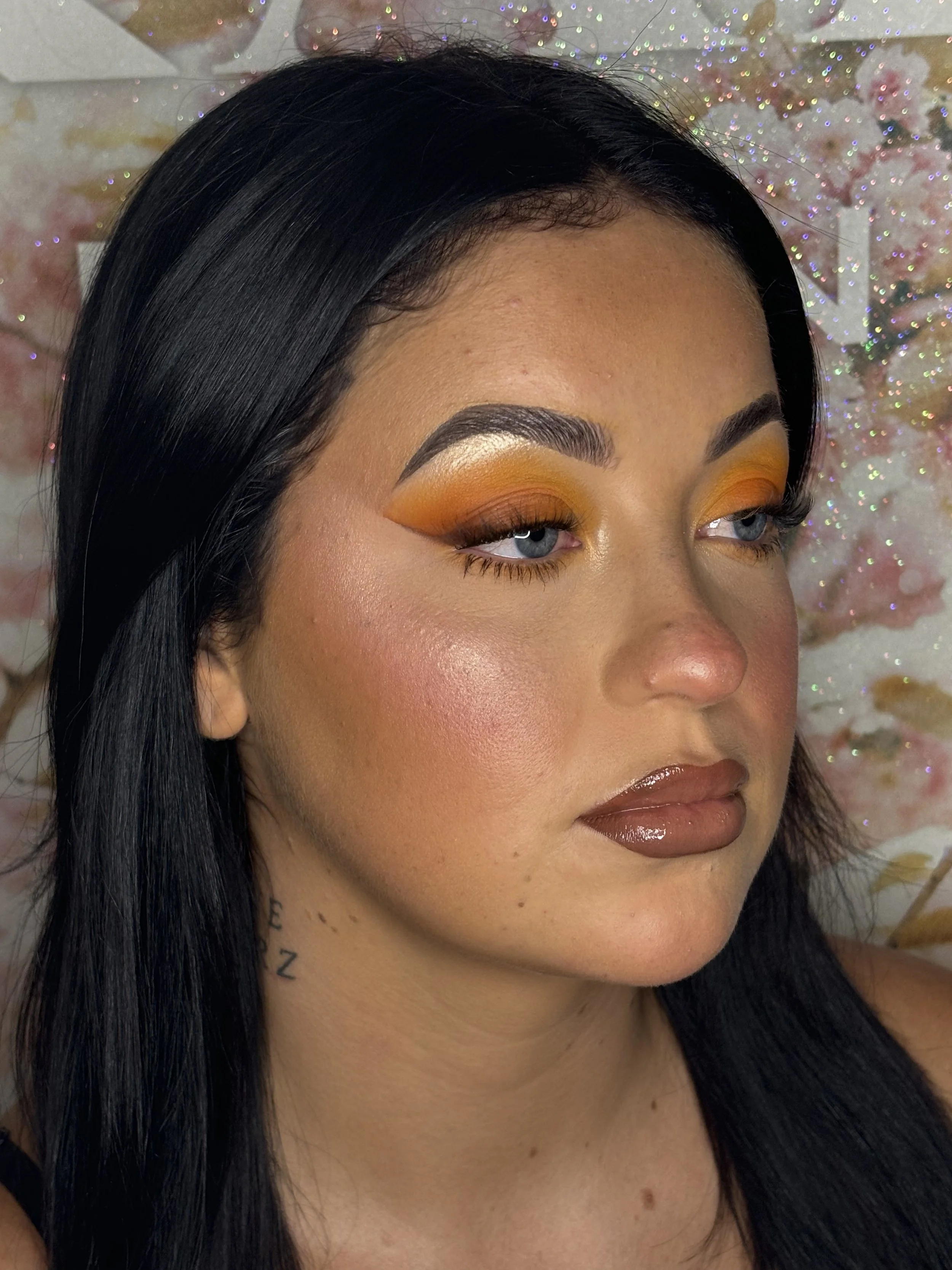 A woman with long black hair and makeup, including orange eyeshadow, glossy brown lipstick, and highlighter, against a glittery floral background.