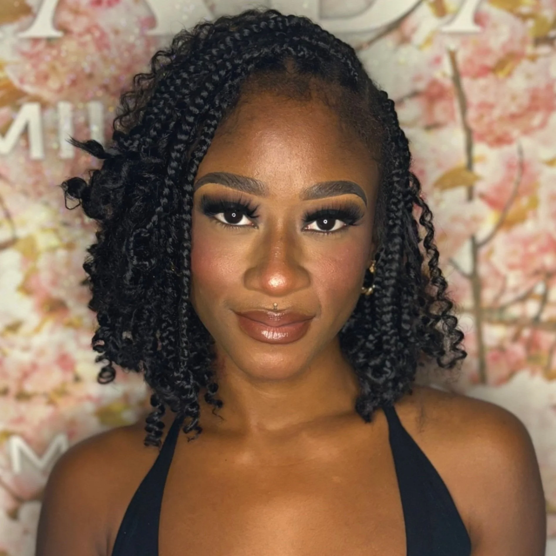 Close-up portrait of a woman with curly black hair, wearing makeup including bold eyebrows, eyeliner, and lipstick, smiling slightly in front of a floral background.