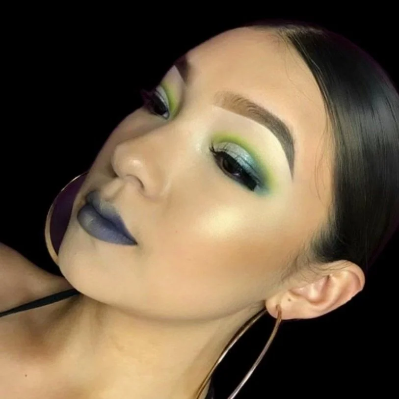 Close-up of a woman with bold, colorful makeup, including blue lipstick, green and yellow eyeshadow, and well-defined eyebrows, with long dark hair and large hoop earrings.