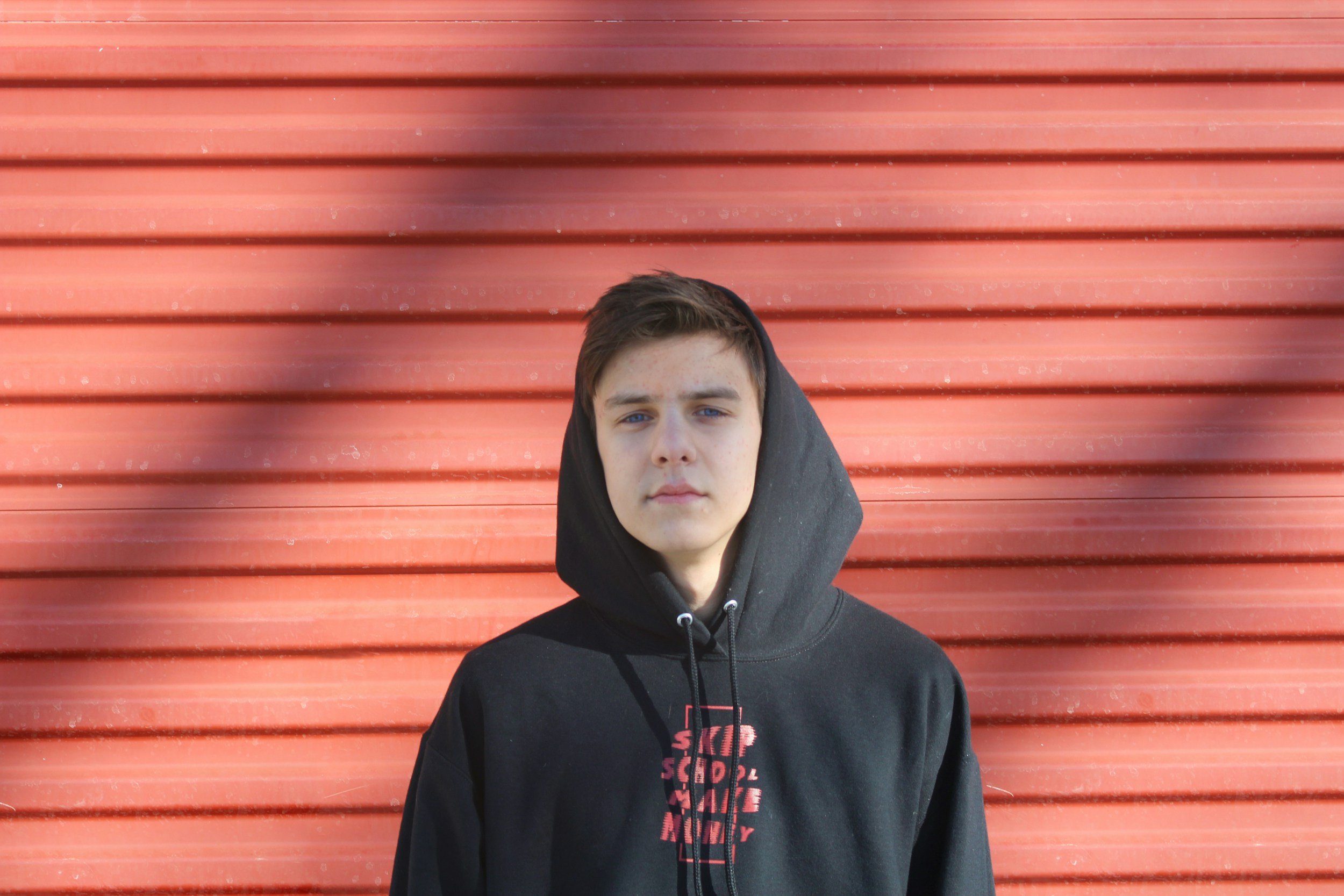 A teenage boy with brown hair and blue eyes wearing a black hoodie stands in front of a red metal wall with horizontal ridges.