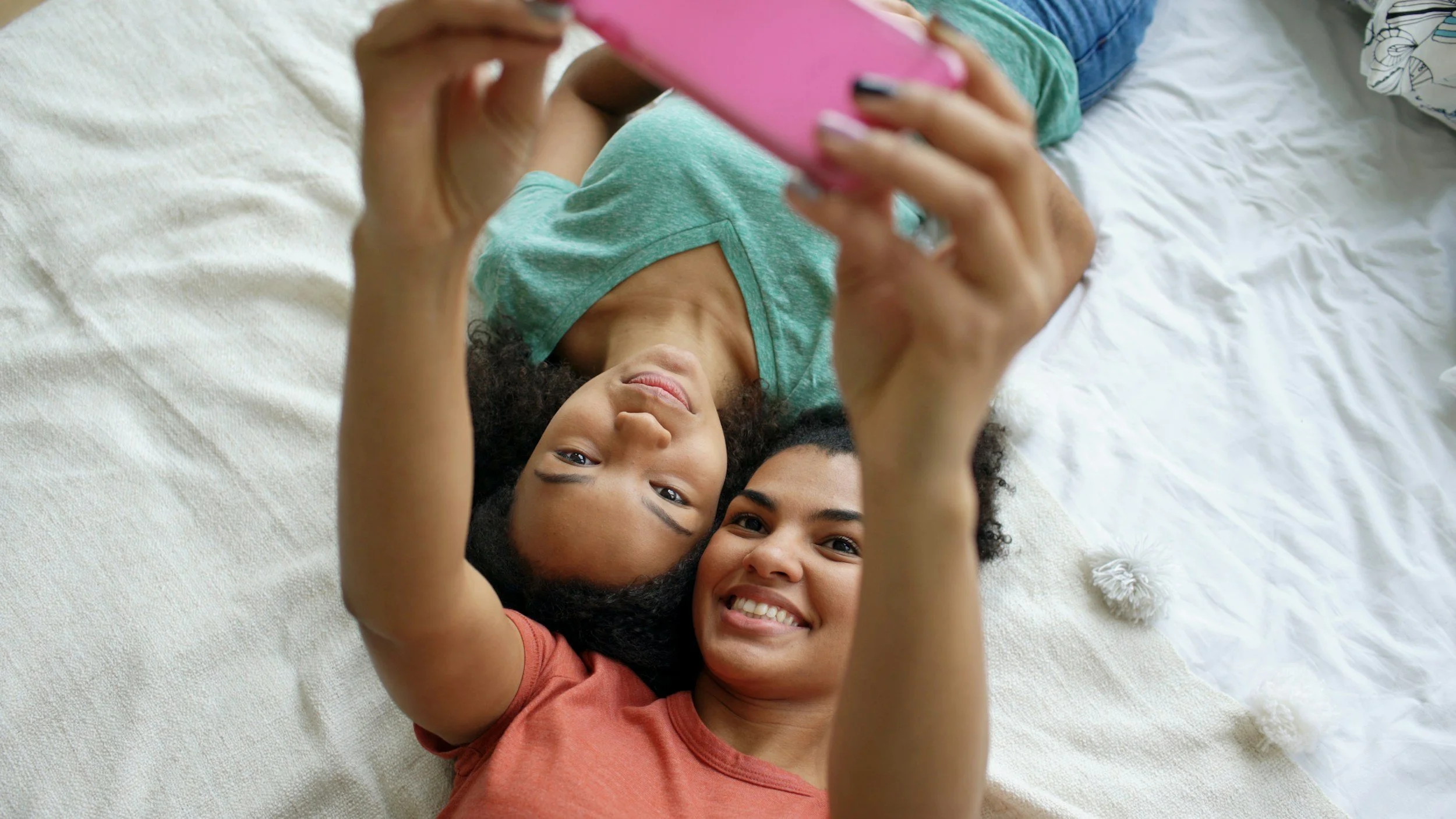 Two smiling young girls lying on a bed and taking a selfie together.