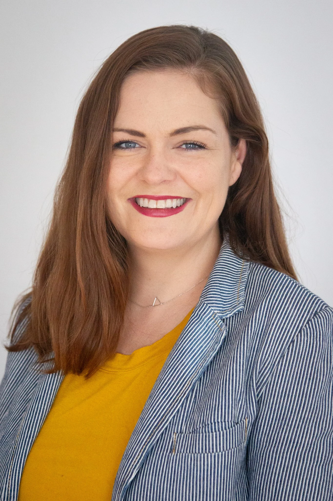 Close-up portrait of a young woman with long, reddish-brown hair, blue eyes, wearing a striped blazer over a yellow top, and smiling with visible teeth.