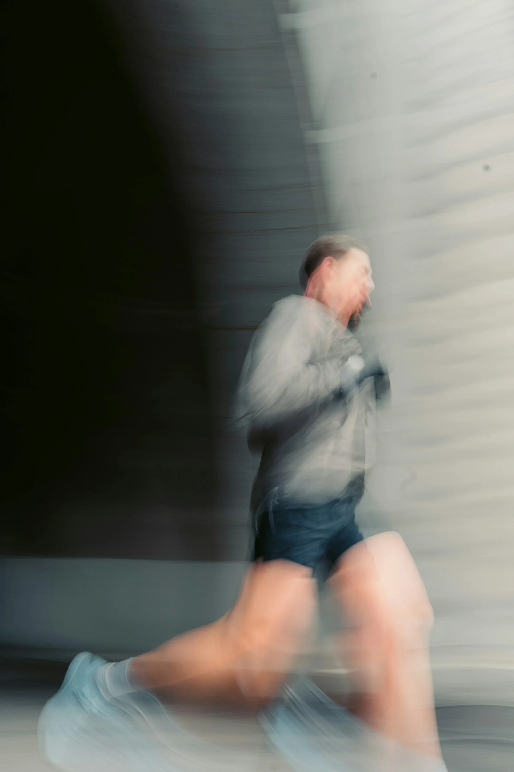 A person running indoors, wearing a gray jacket, black shorts, and white shoes, captured with motion blur.