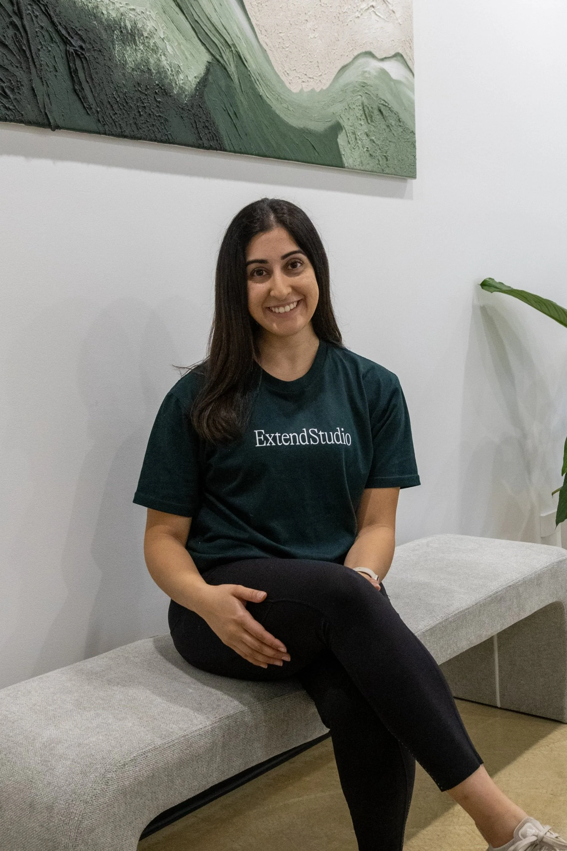 A woman with long dark hair sitting on a light gray bench, smiling, wearing a dark green ExtendStudio T-shirt, black pants, and beige sneakers, in an indoor setting with white walls, green abstract artwork, and a large potted plant.