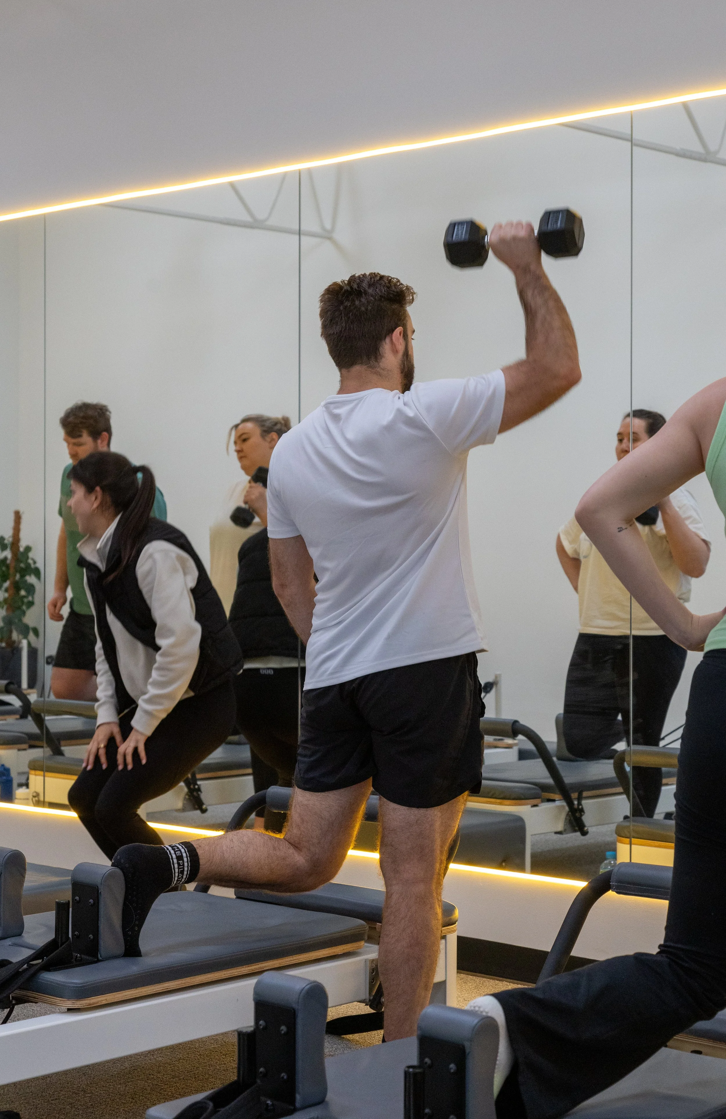 A man is sitting cross-legged on a Pilates reformer machine, holding a ring exercise equipment, and smiling at a trainer who is standing beside him, also smiling. The setting appears to be a Pilates or fitness studio.