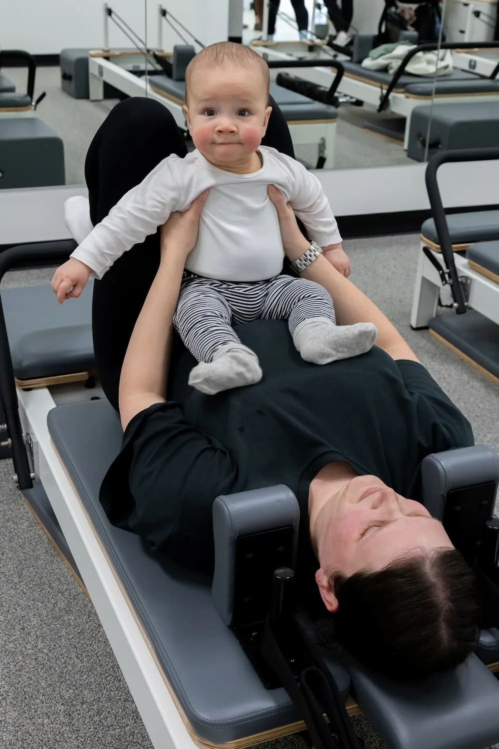 A baby sits on a person's stomach on a medical examination table, with the person lying down and the baby looking at the camera in a medical or therapy facility.