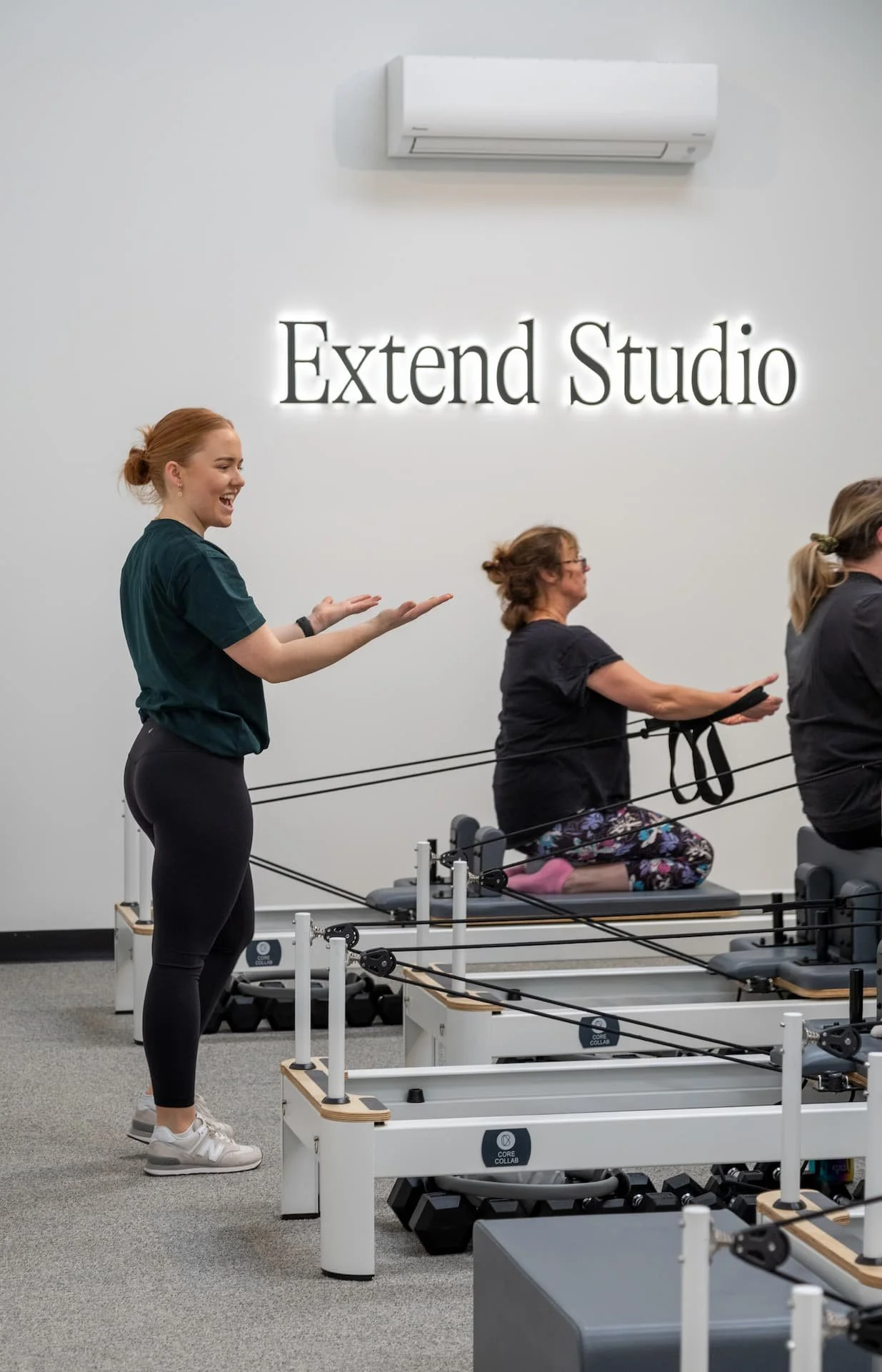 A fitness instructor leading a Pilates class in a studio with a white wall and a sign that reads 'Extend Studio'. Two women are exercising on Pilates reformer machines, one sitting and the other kneeling. All participants are wearing workout clothes.