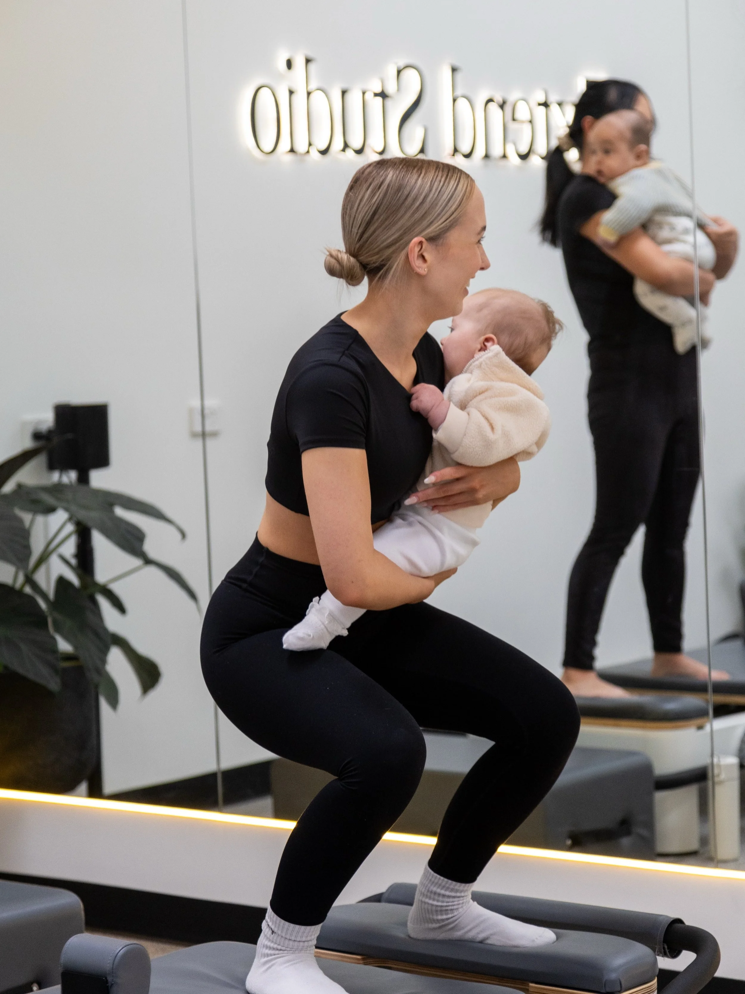 A woman in black workout clothes is on an exercise step, holding a baby in beige clothing while performing a squat. In the background, another woman is holding a child, standing on a similar exercise step in front of a mirror with a logo that reads 'studio'.