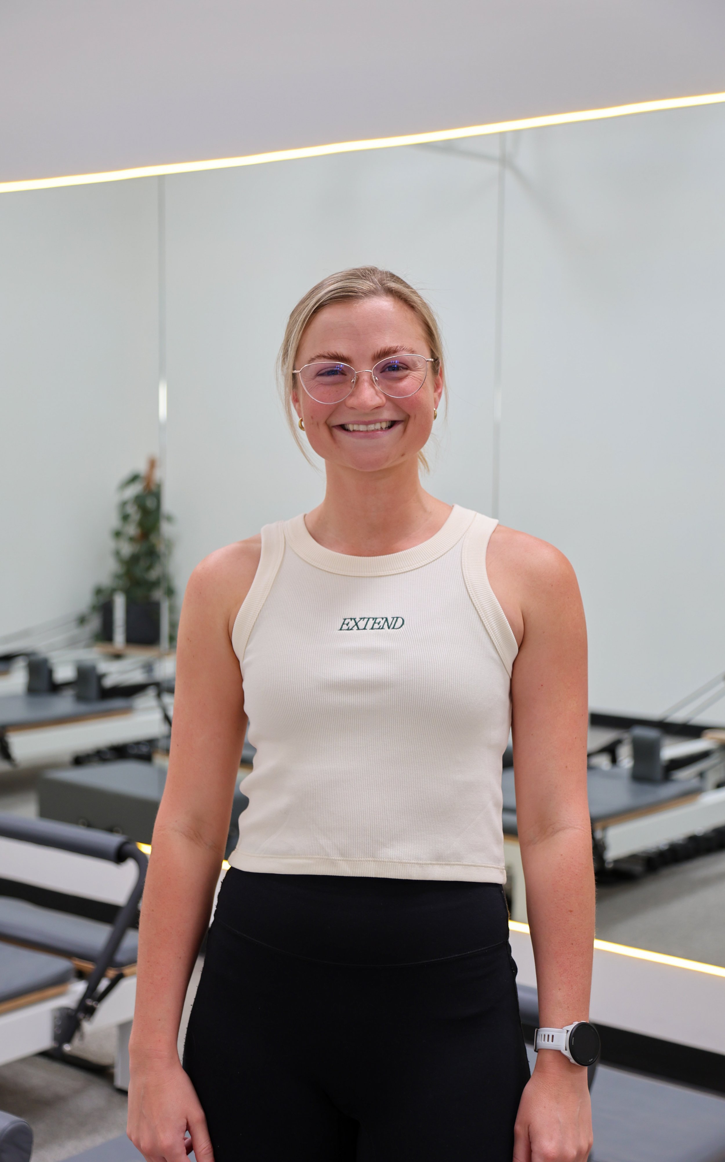 A smiling woman in a sleeveless gray workout top standing in a fitness studio with a mirror behind her.