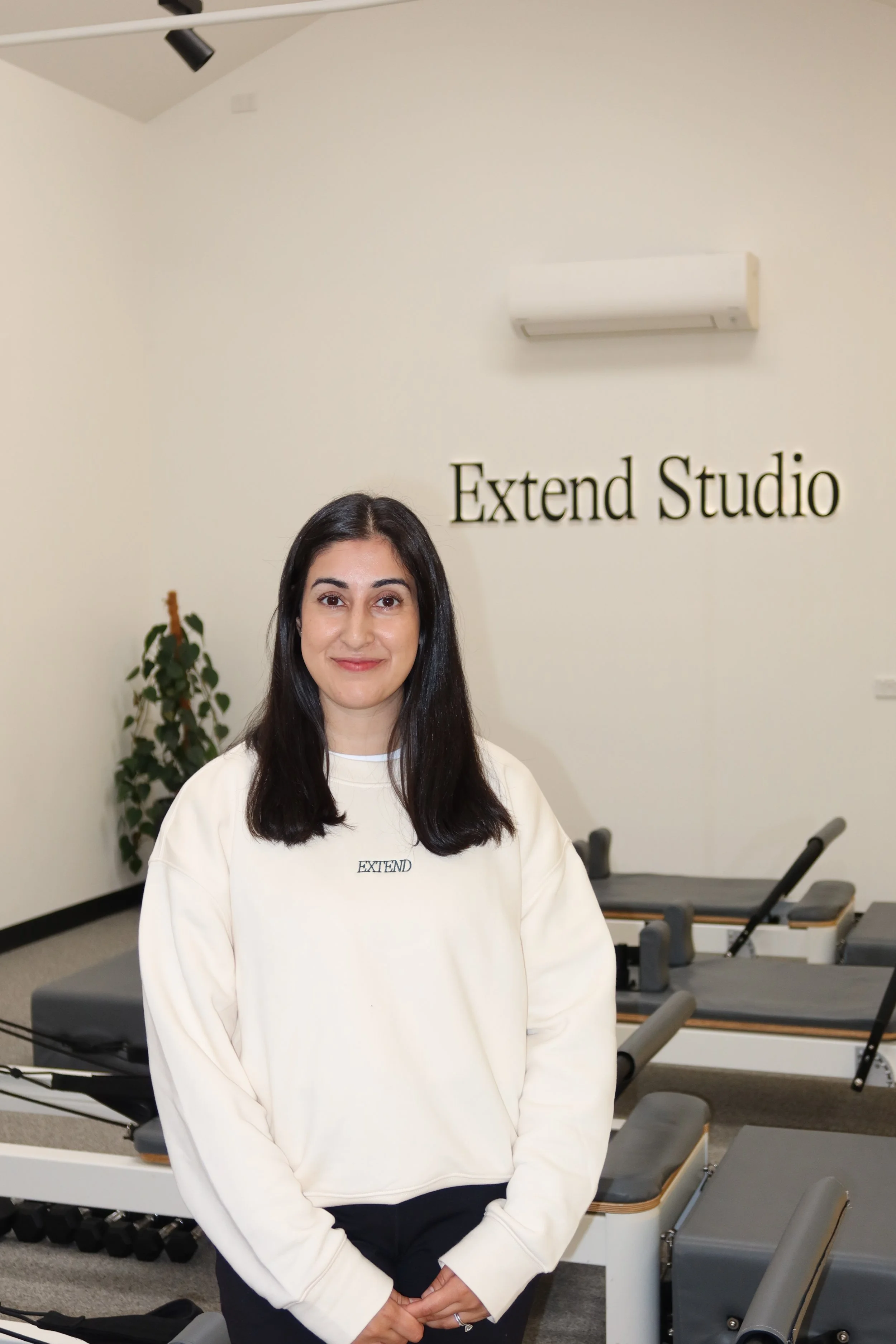 A woman with long dark hair sitting on a light gray bench, smiling, wearing a dark green ExtendStudio T-shirt, black pants, and beige sneakers, in an indoor setting with white walls, green abstract artwork, and a large potted plant.