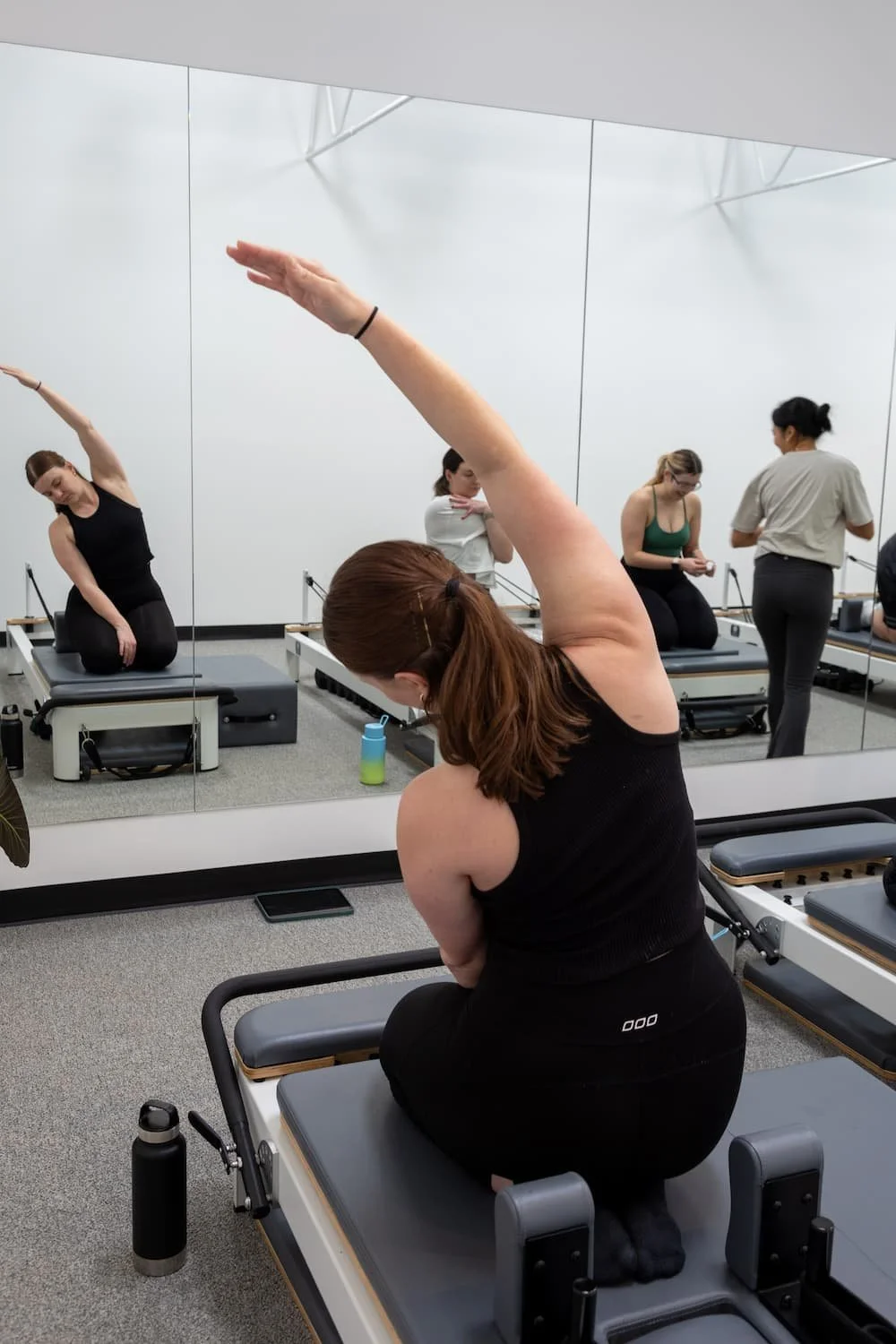 A woman practicing yoga in a Pilates studio, kneeling on a reformer machine with one arm extended upward, while looking into a large mirror that reflects her and other people in the studio.