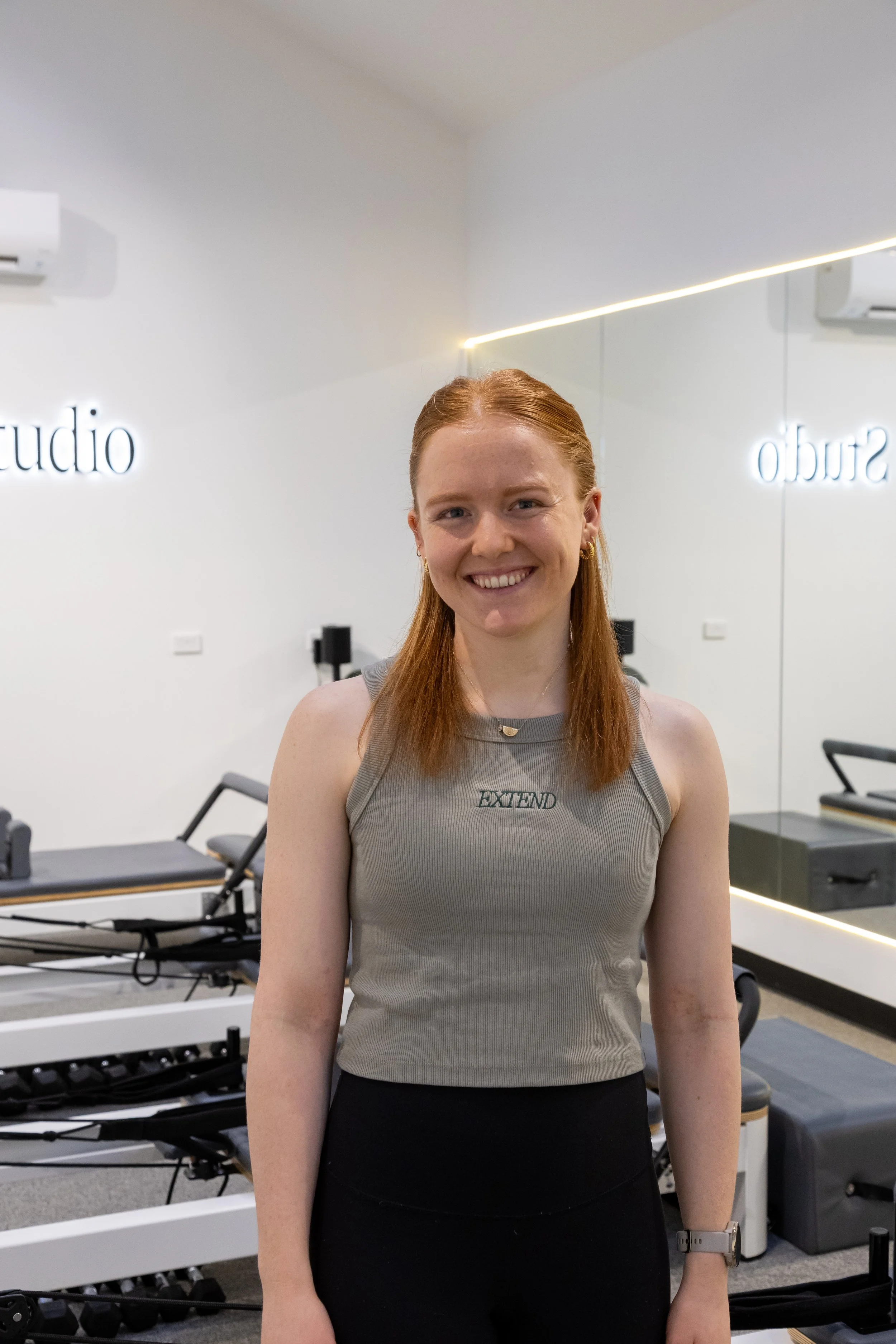 A smiling red-haired woman sitting on a light gray bench with her hands clasped, wearing a white ExtendStudio sweatshirt, black leggings, and white sneakers, in a modern, well-lit room with abstract green artwork and large plants.