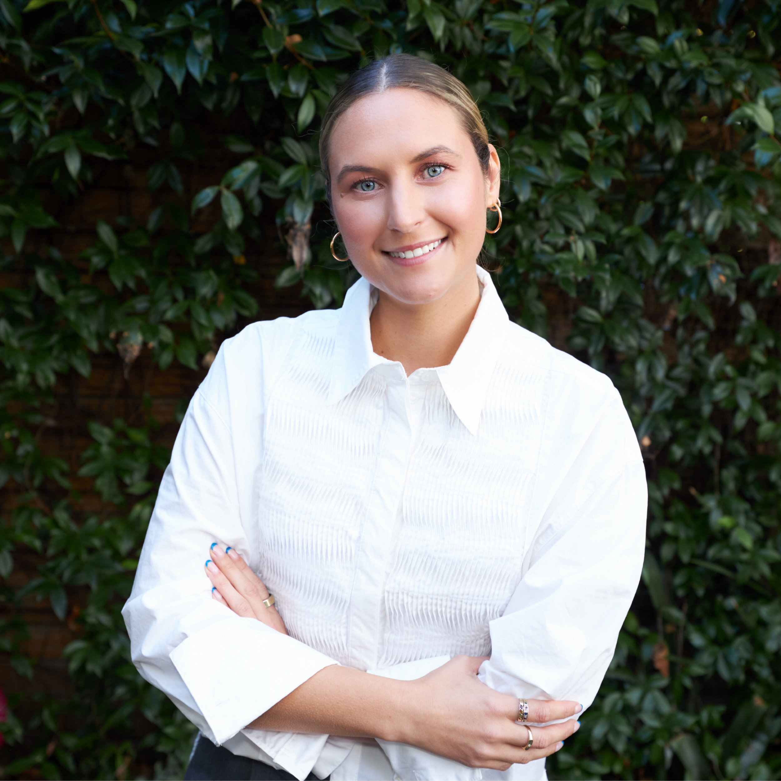 Melbourne naturopath, Jessica Diakoumakos wearing a white ruffled shirt and standing in front of a wall of vines, smiling