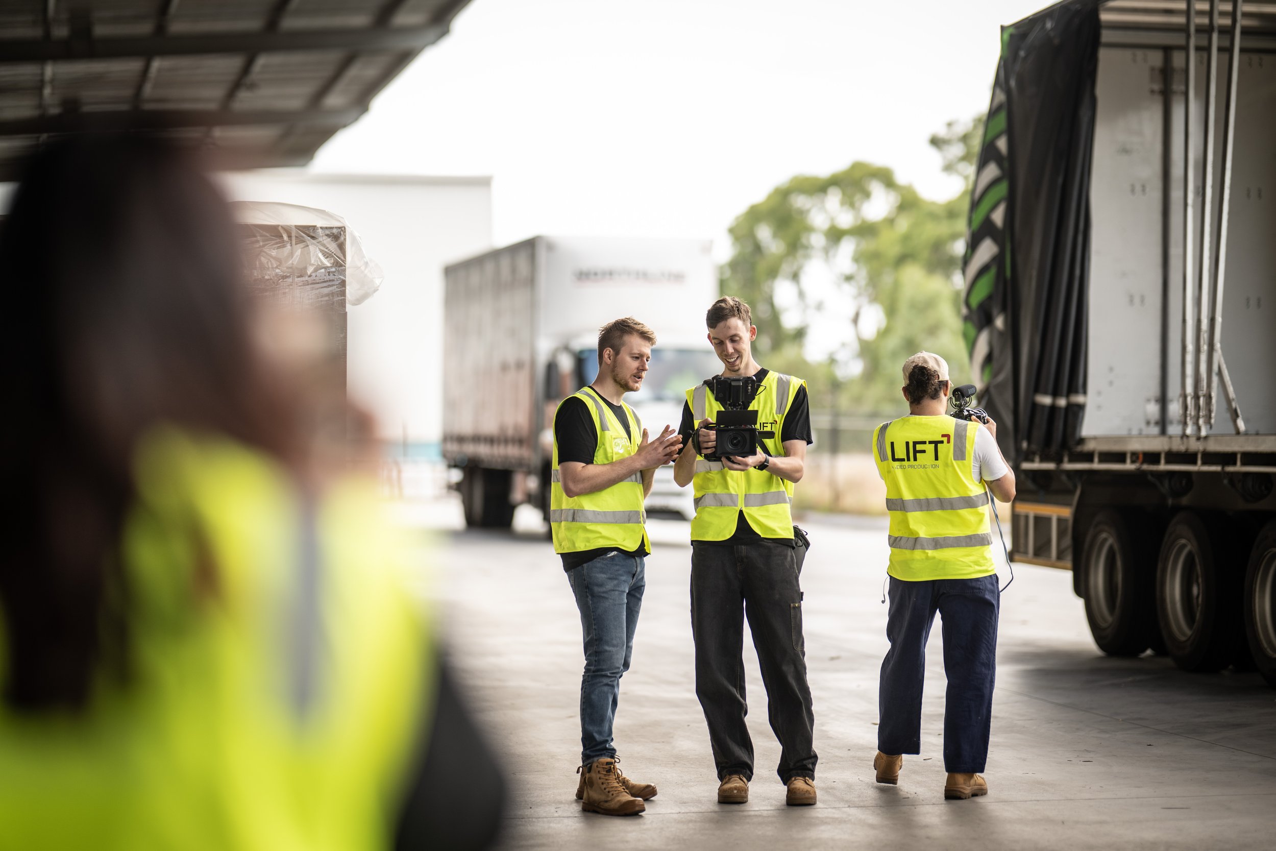 Three construction workers in yellow safety vests standing under a truck, discussing a camera or device, with two additional workers in the background, all outdoors.