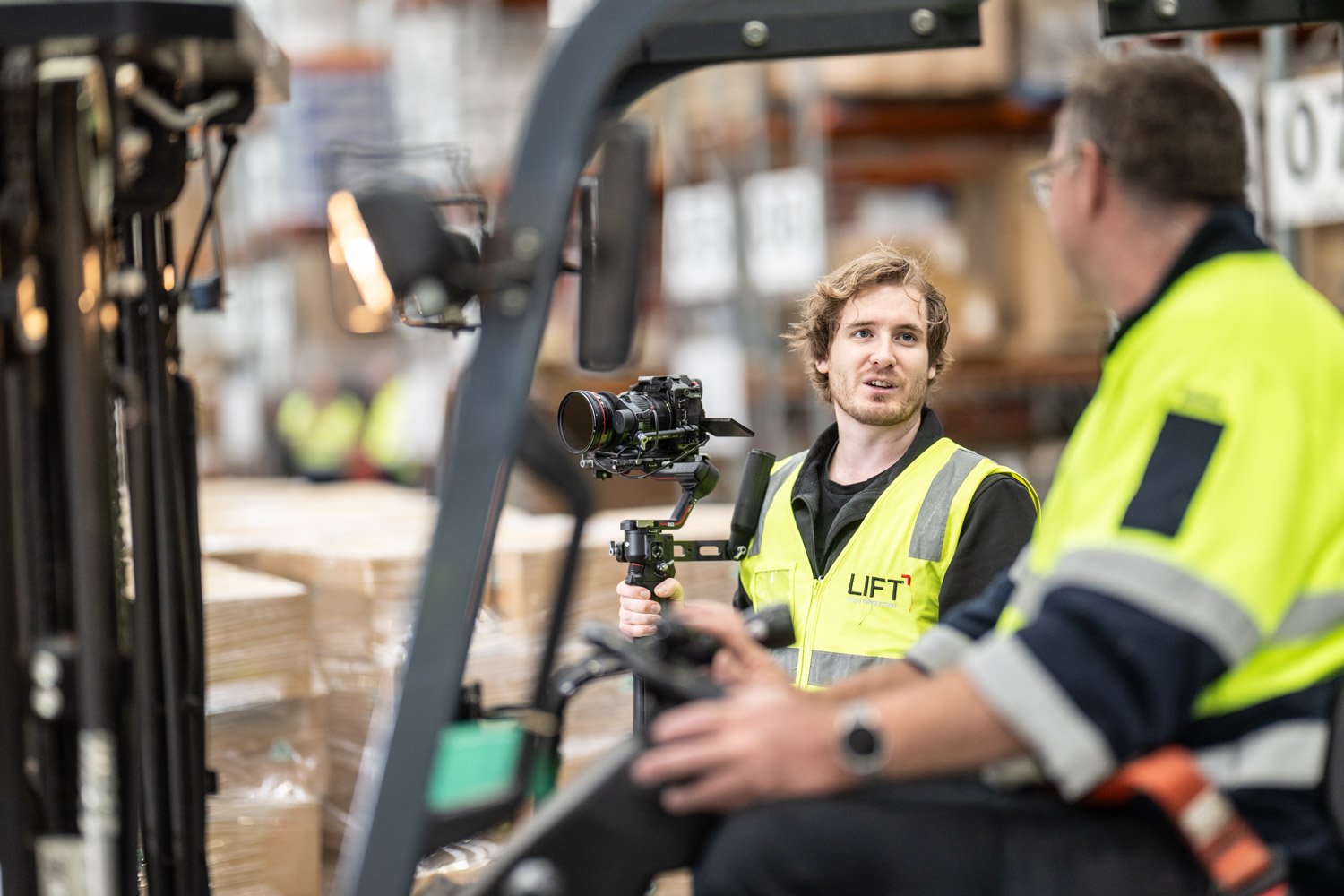 Two warehouse workers in high-visibility jackets working with a forklift. One worker is talking, and the other is seated and holding a remote control device.