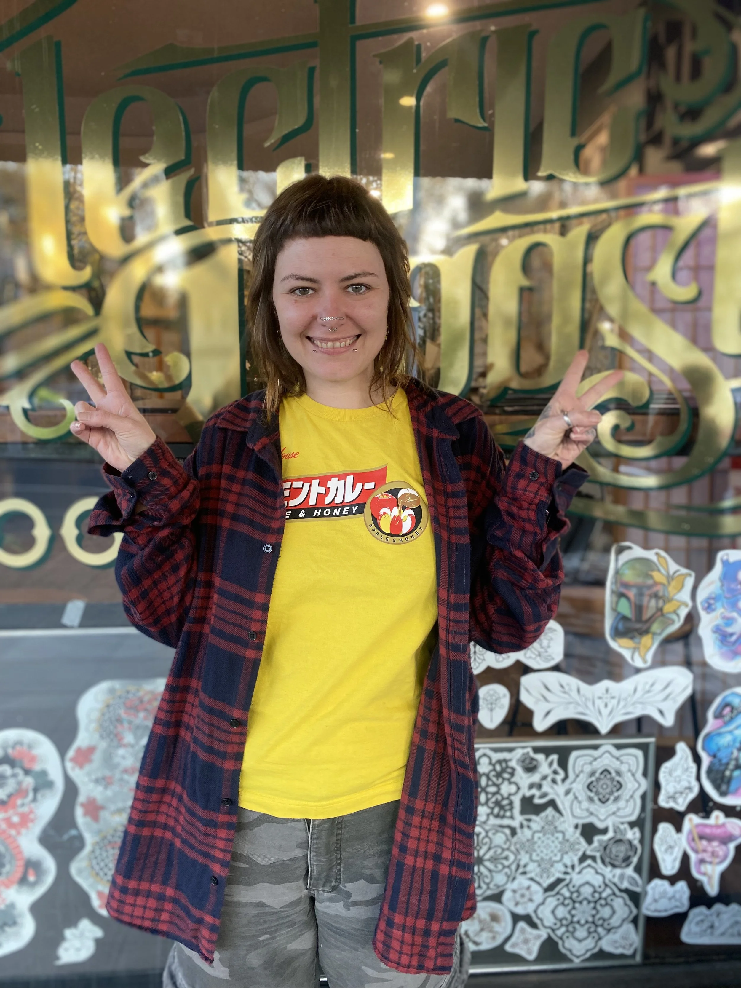 A woman with short brown hair, wearing a yellow T-shirt with a Japanese-style graphic and text, a red and black checkered flannel shirt, and camouflage shorts, standing in front of a window with gold lettering and displaying various art stickers.