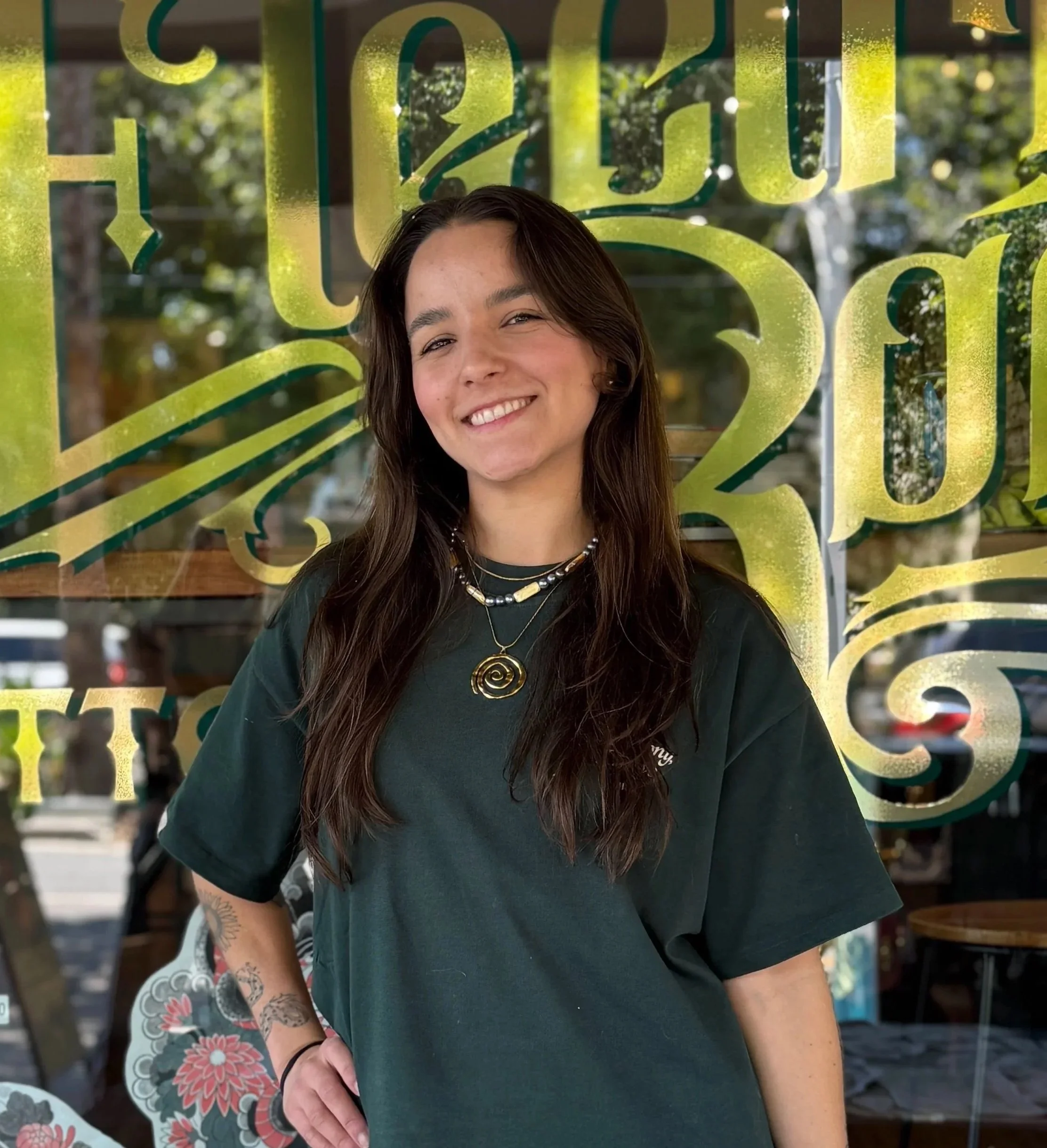 A young man with short hair, beard, and gauges standing in front of a window with gold lettering. He is wearing a black T-shirt with a colorful dragon graphic and the text 'Devil Apparel.' He has tattoos on his left arm.