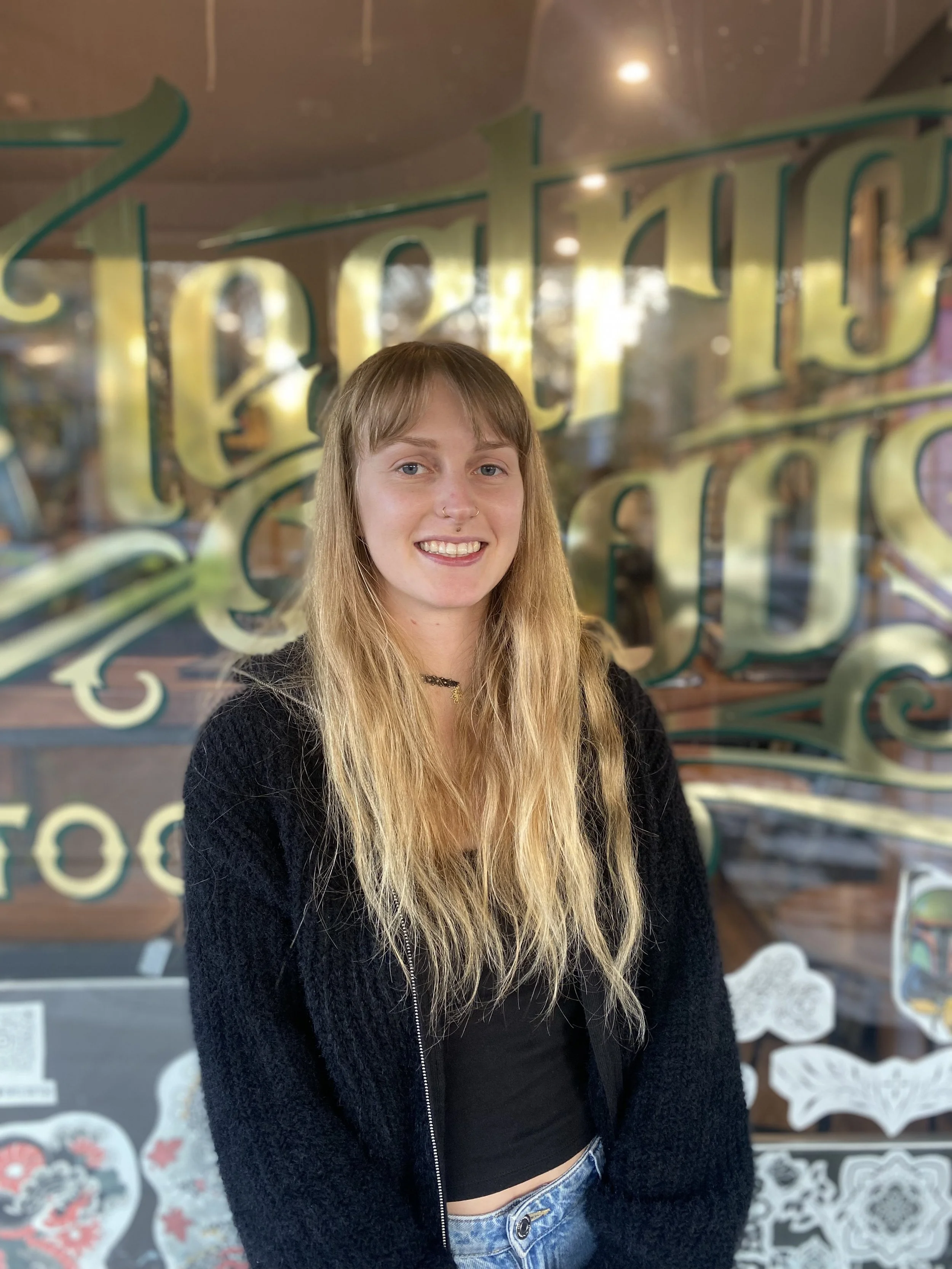 Young woman with long blonde hair, black top, black zip-up jacket, and blue jeans stands in front of a window with decorative writing and artwork.