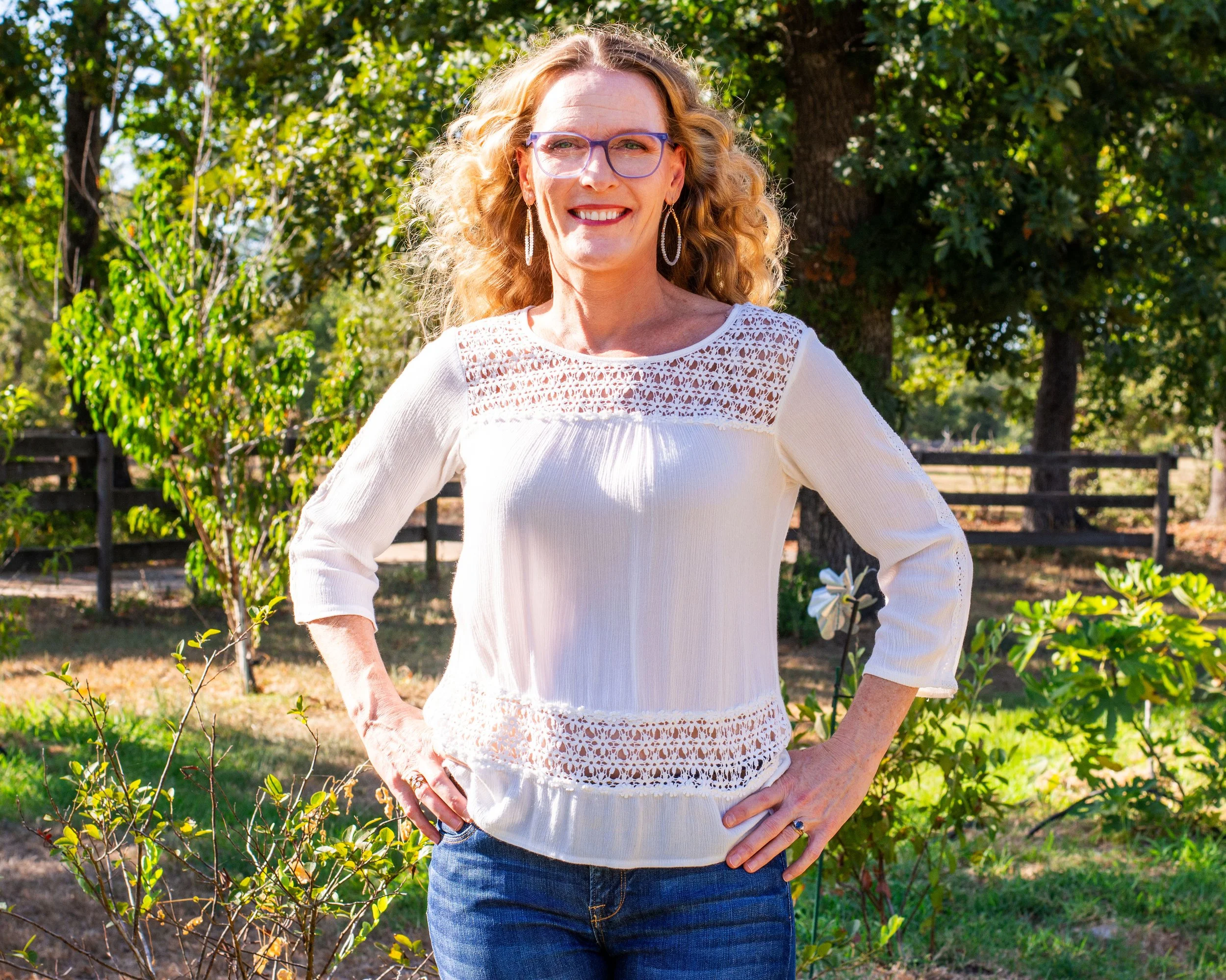 A woman with curly blonde hair and glasses standing outdoors in a park with trees and a fence, smiling and posing with her hands on her hips.