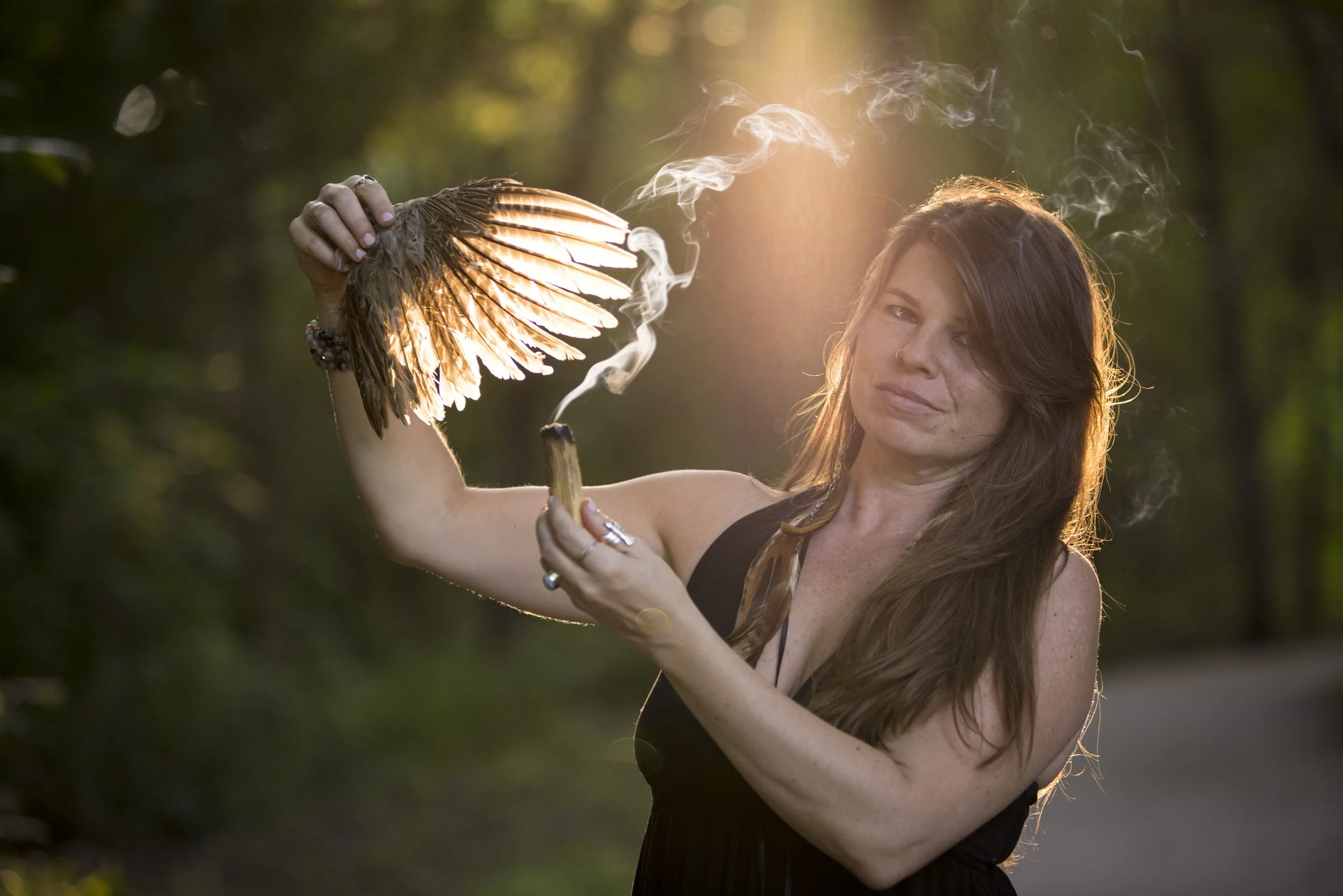 A woman with long brown hair holding a bird with glowing wings and smoke rising from it during sunset in a wooded area.