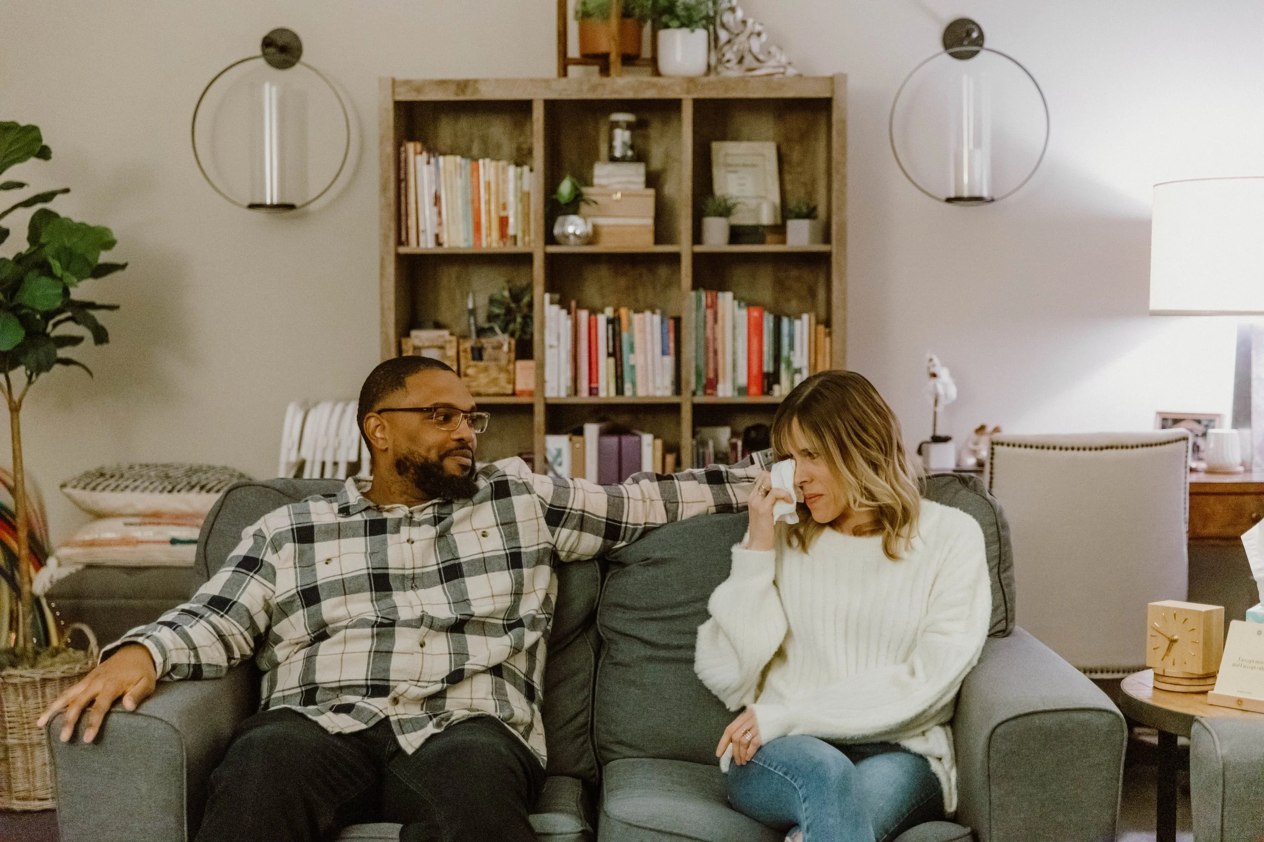 A biracial couple on a couch during a couples intensive therapy session in Chino, California. The wife wipes away a tear during an emotional moment. A therapist trained in marriage intensives supports the process of reconnecting.