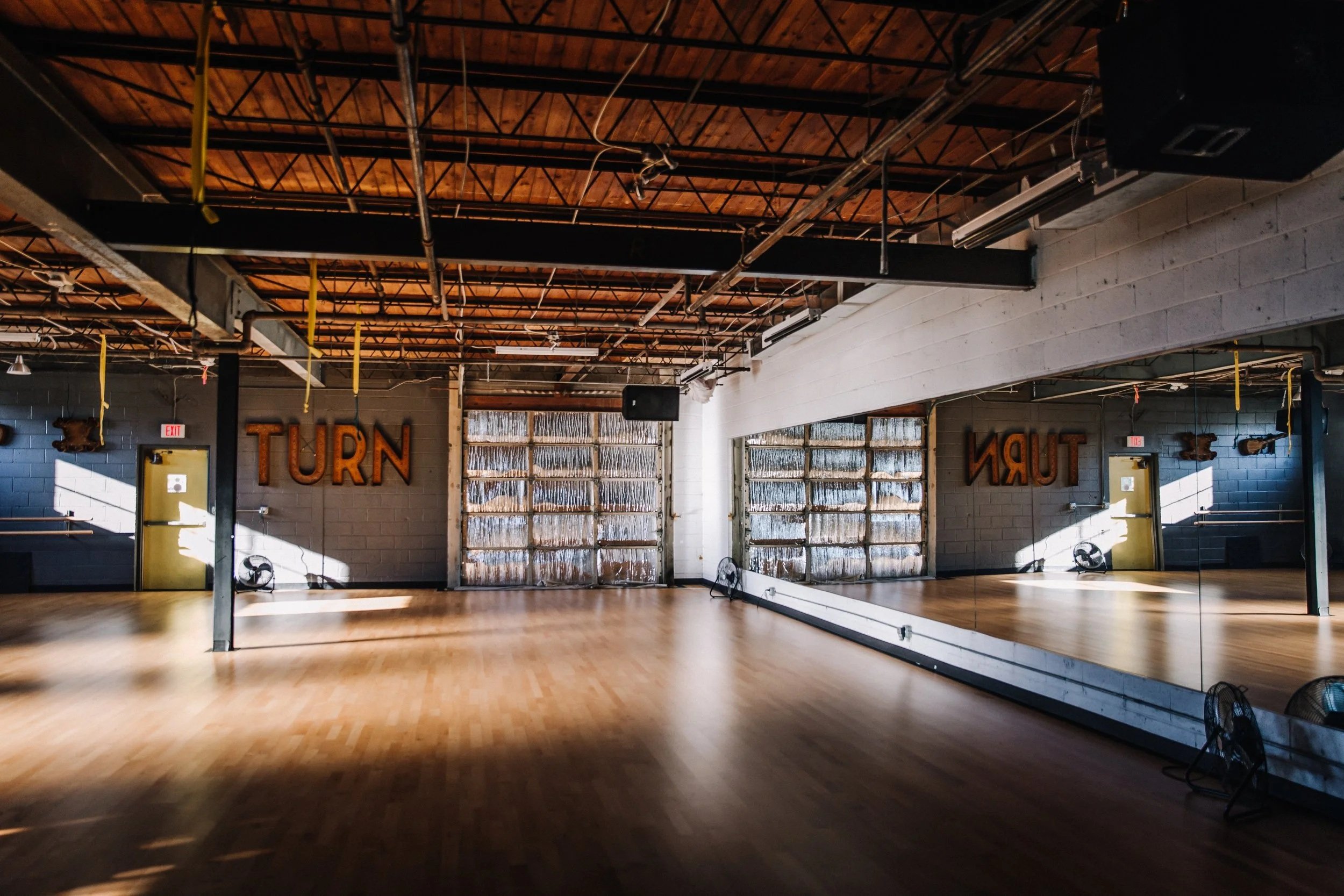 Dance studio with wooden floors, large mirror, and the word 'TURN' on the wall. The studio is lit by natural light from windows, and has industrial-style ceiling with exposed pipes and lighting.