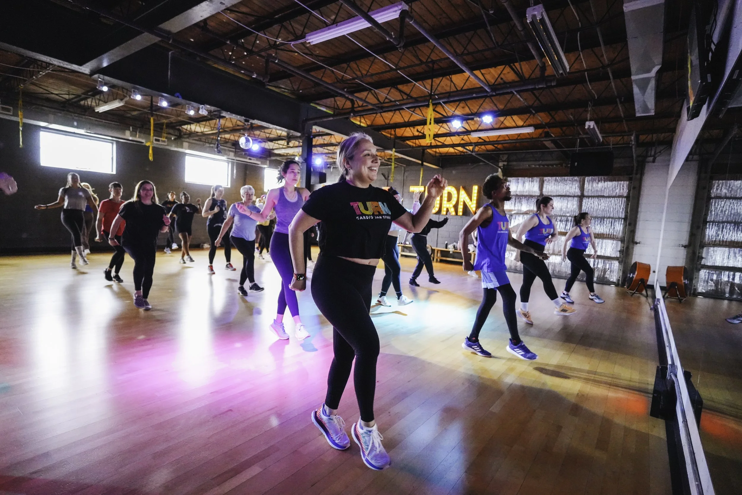 Group of women participating in a dance or fitness class in a studio with wooden floors and high ceilings, colorful lights, and a neon sign in the background.