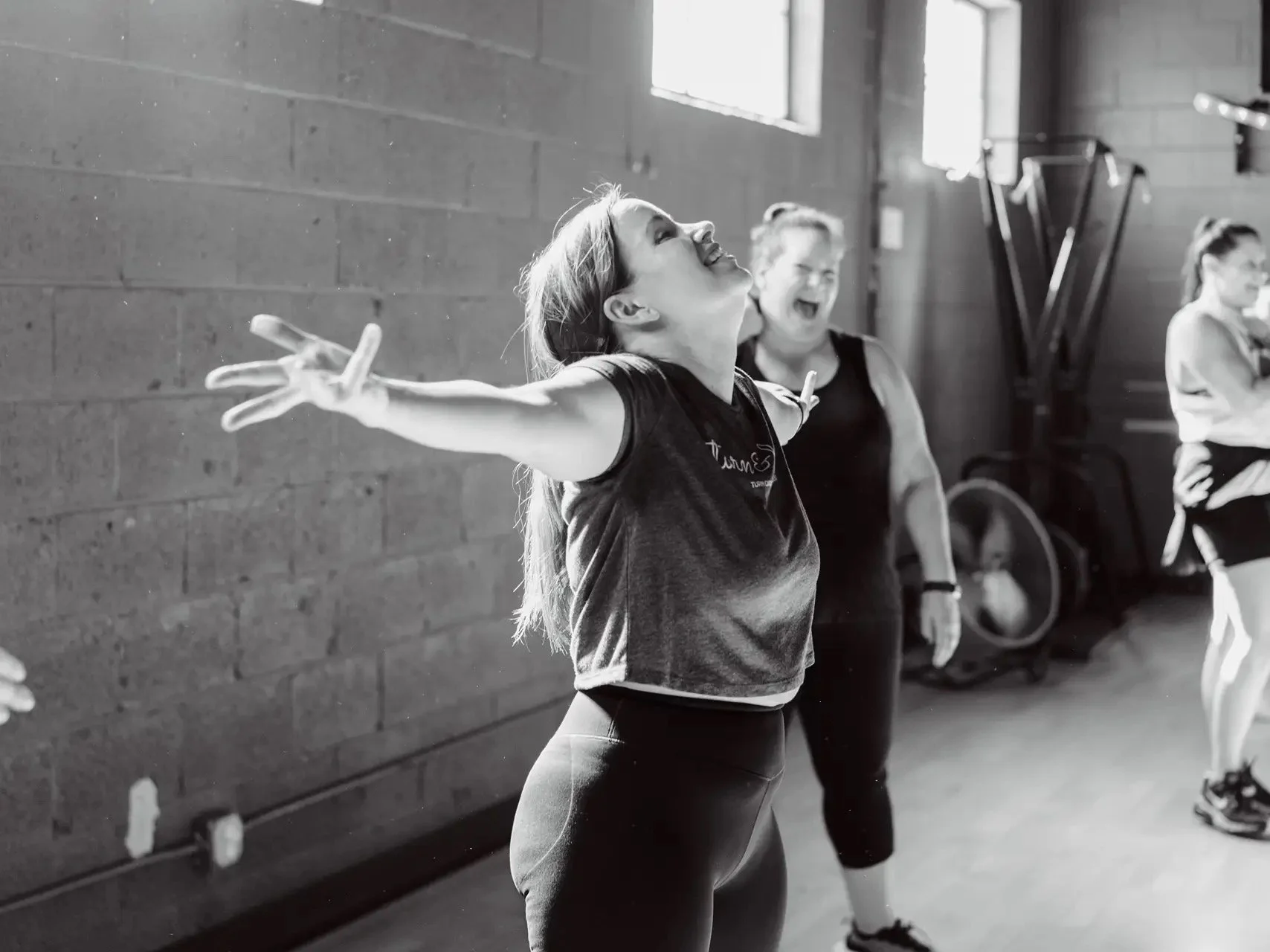 Women in a gym class celebrating with arms outstretched, smiling, with others in the background.
