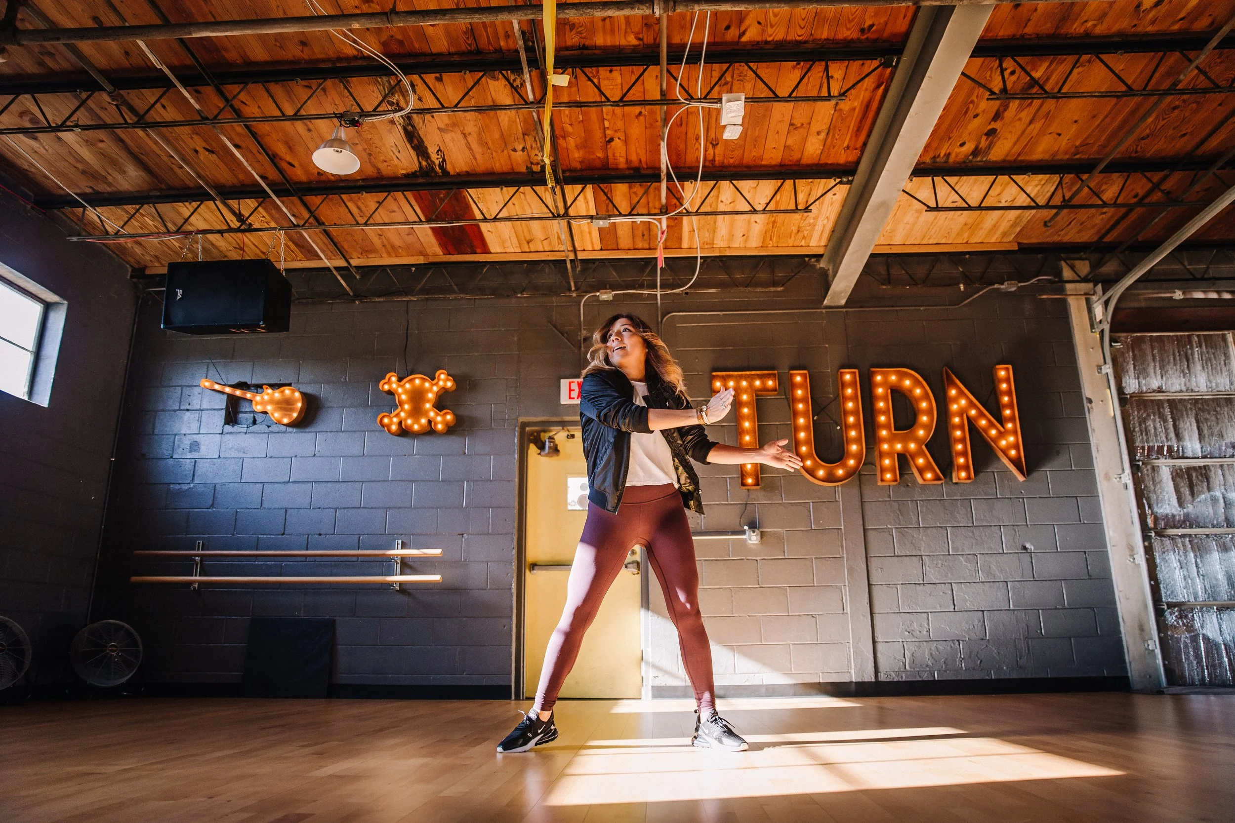 A woman dancing in an industrial-style dance studio with a black brick wall and wooden ceiling, illuminated by warm bulbs spelling 'TURN' and decorative lights on the wall.