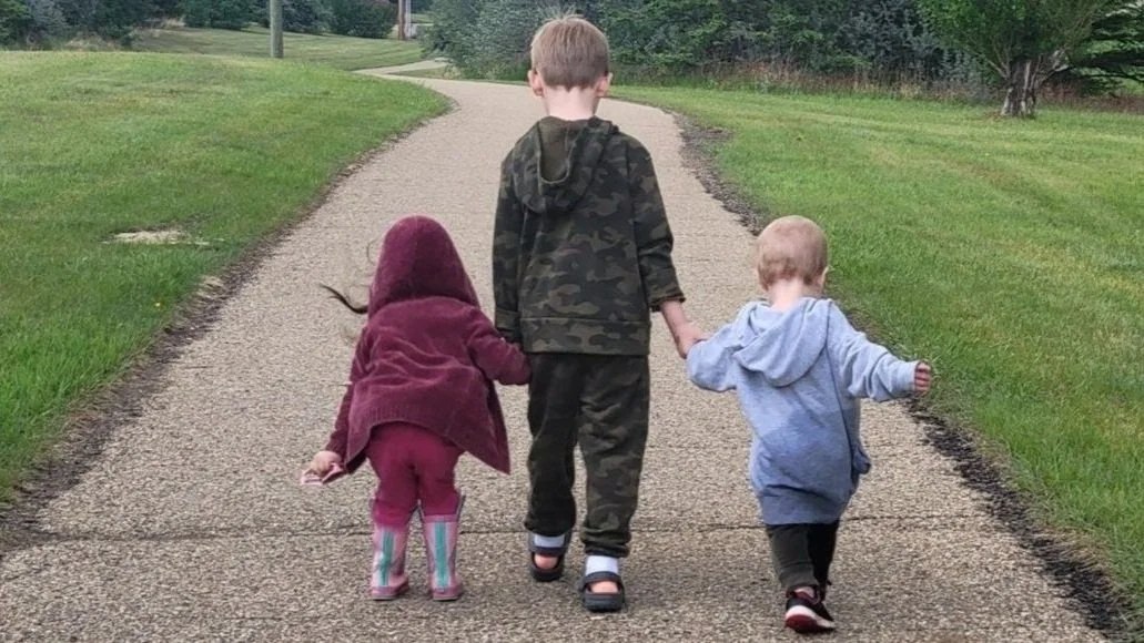 Three children walking on a path outdoors, with two holding hands and connected to the middle child, who is in the center of the image, walking away from the camera.