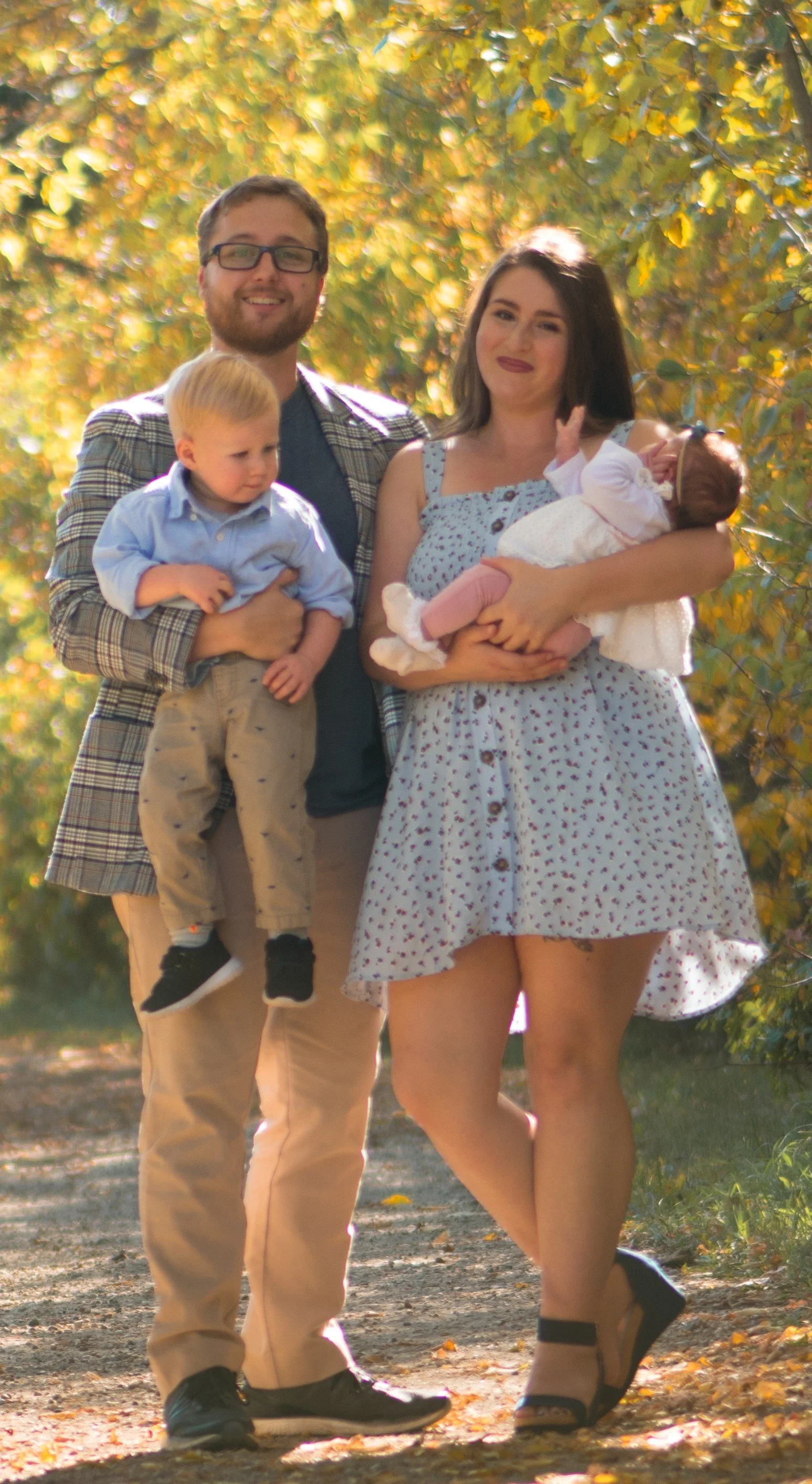 A family of four standing outdoors on a sunny autumn day with yellow and green leaves in the background. The man is holding a young boy, and the woman is holding a baby.