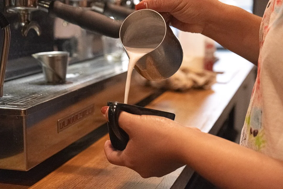 Barista pouring steamed milk into a black coffee cup at a coffee shop counter.