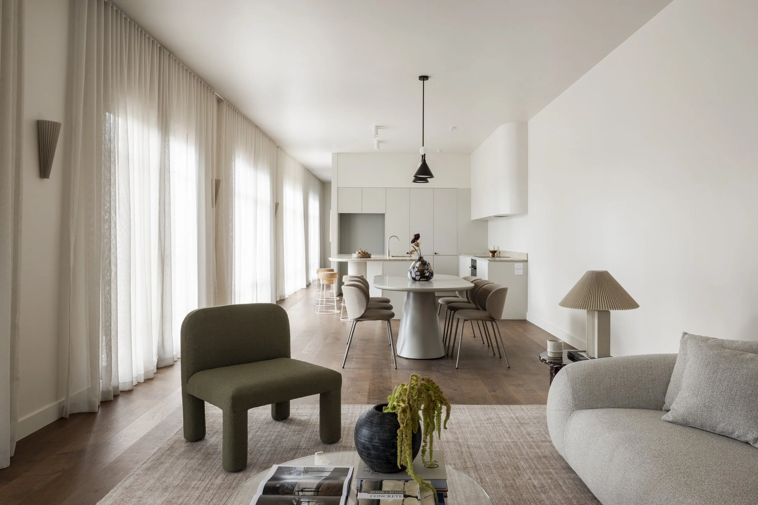 Modern, minimalist living room and kitchen with white walls, large sheer curtains, and wooden flooring. The living room has a light gray curved sofa, a dark green chair, and a coffee table with books and a black vase with green plants. The dining area features a round table with beige chairs, and the kitchen has white cabinetry and a marble countertop. There is a black pendant light hanging above the dining table and table lamps in the living area.
