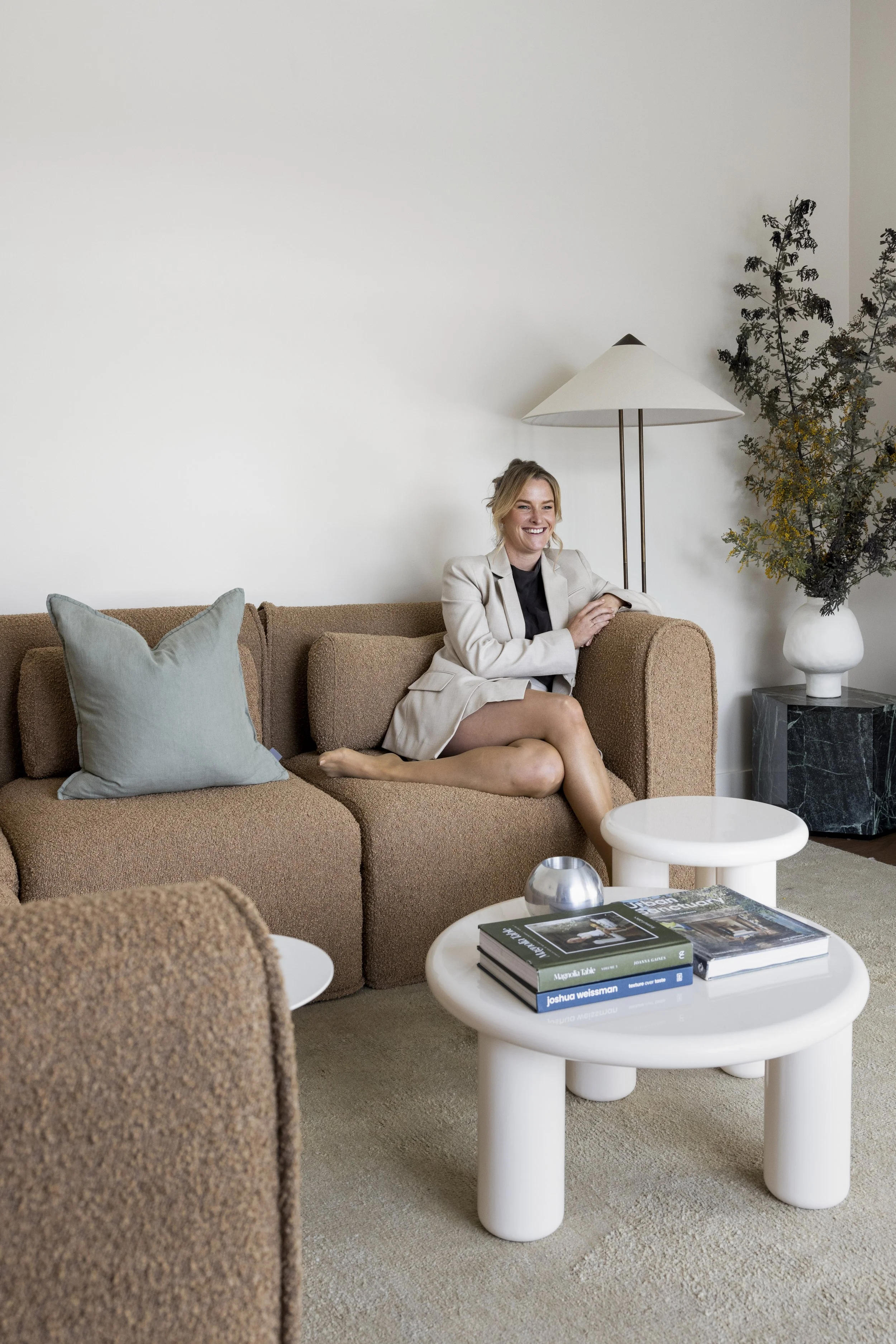 A woman in a beige blazer sitting on a brown sofa, smiling, in a modern living room with a white wall, green pillow, white coffee table with books, a black marble side table, a floor lamp, and a large plant in a white vase.