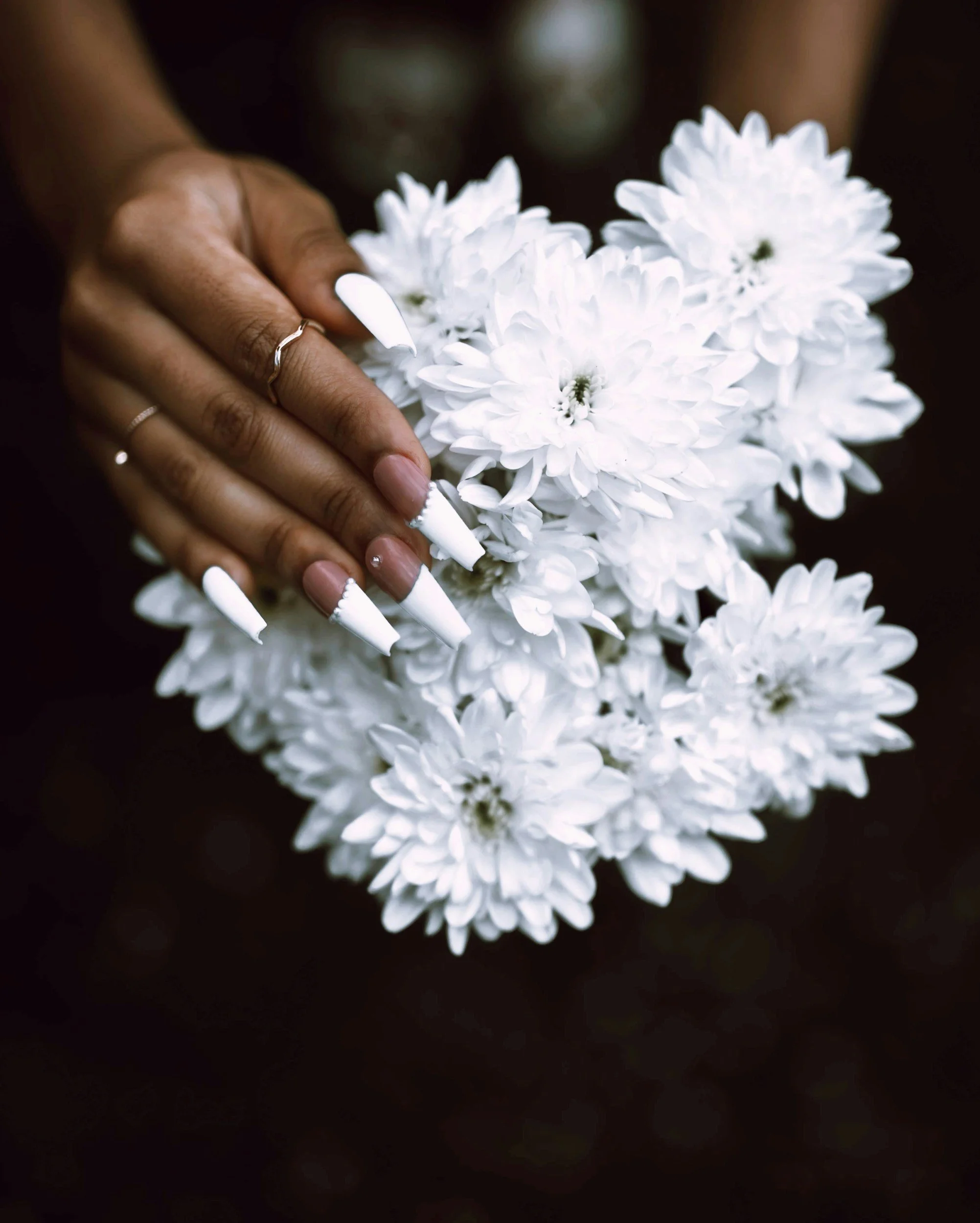 Close-up of a hand with long white nails holding a bouquet of white flowers against a dark background.