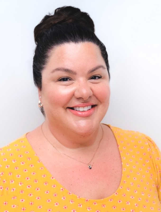A woman with dark hair styled in a bun, smiling, wearing a yellow top with pink and white floral pattern, earrings, and a necklace, against a plain white background.