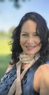 A woman with dark curly hair smiling outdoors with a blurred green park background.